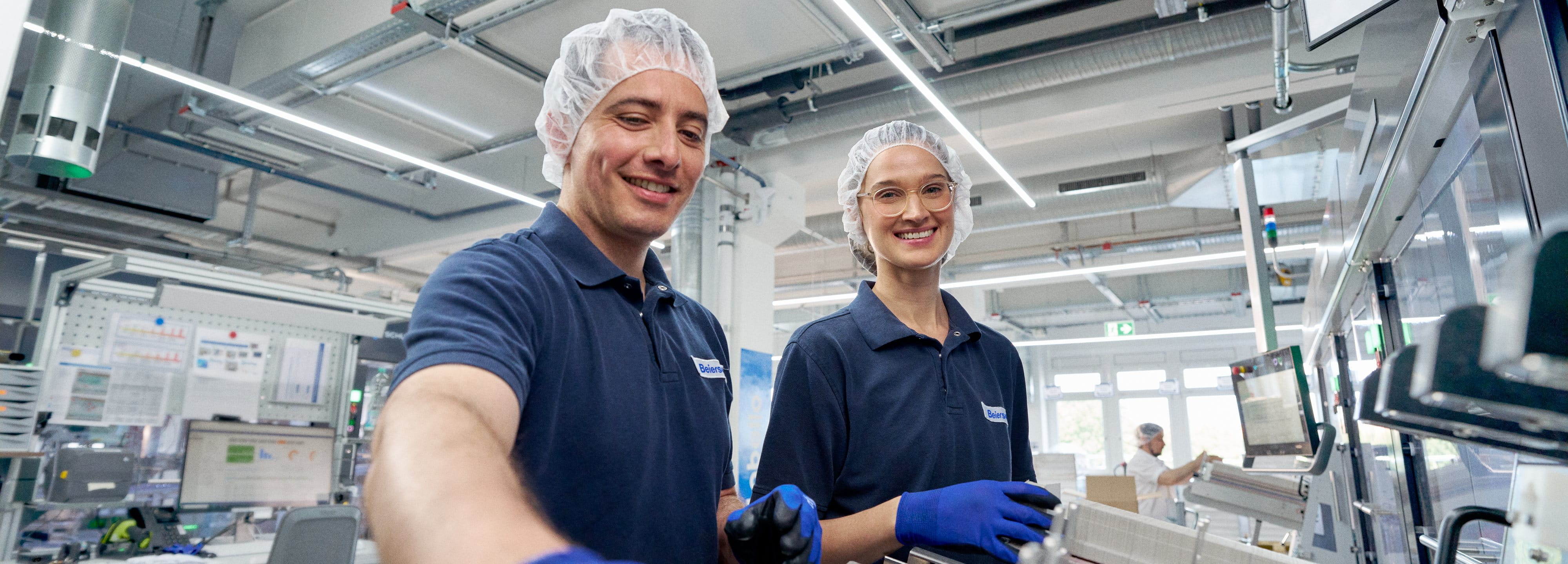 Two factory workers wearing hair nets and blue gloves smiling while operating equipment in a clean industrial production facility.