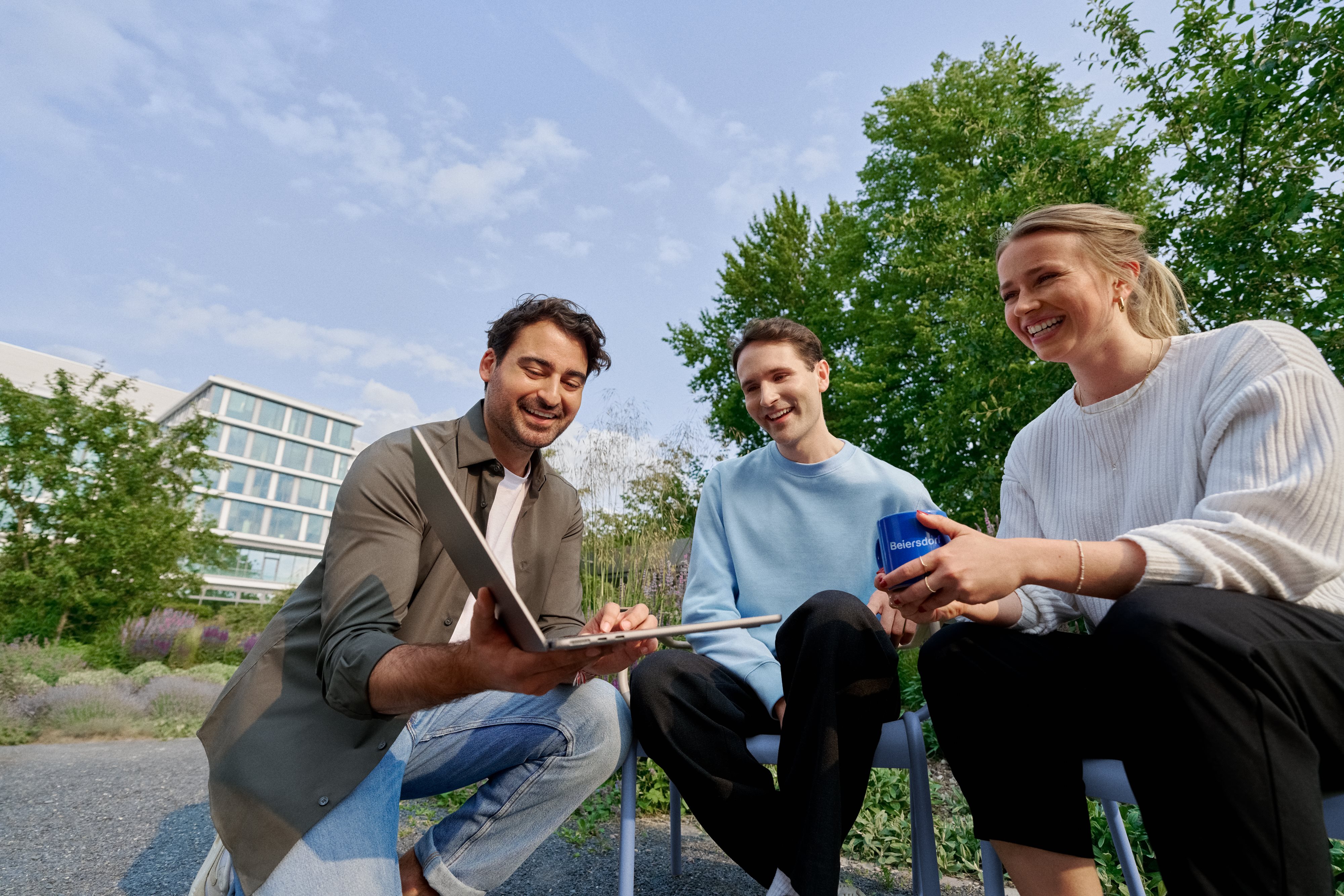 Three smiling coworkers sitting in the campus garden, collaborating on a laptop and enjoying teamwork.