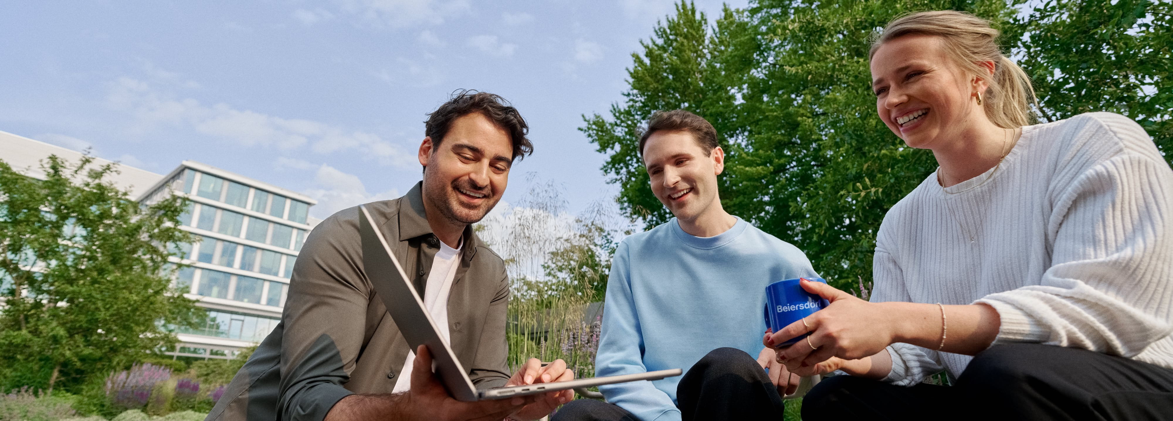 Three smiling coworkers sitting in the campus garden, collaborating on a laptop and enjoying teamwork.