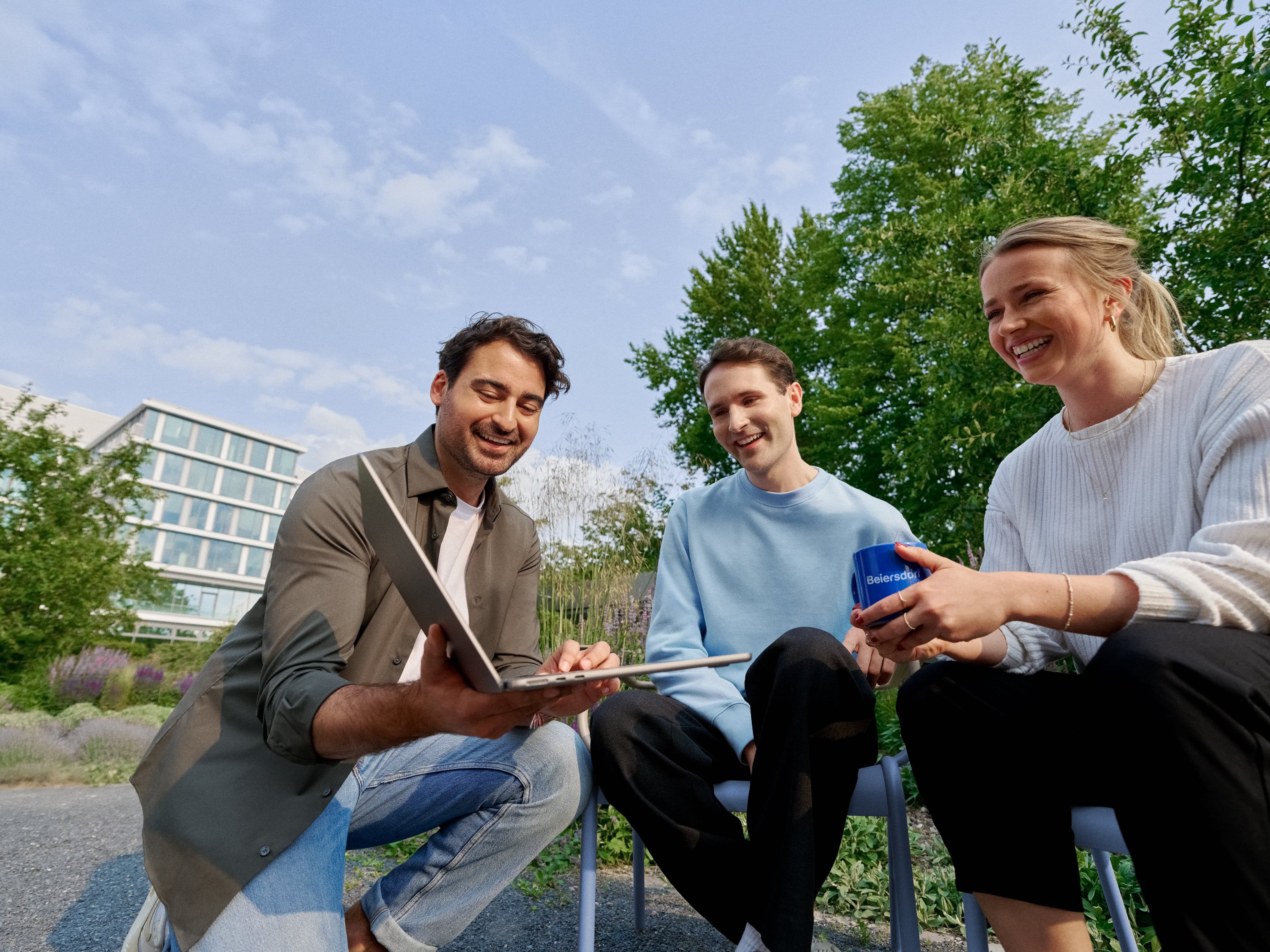 Three smiling coworkers sitting in the campus garden, collaborating on a laptop and enjoying teamwork.