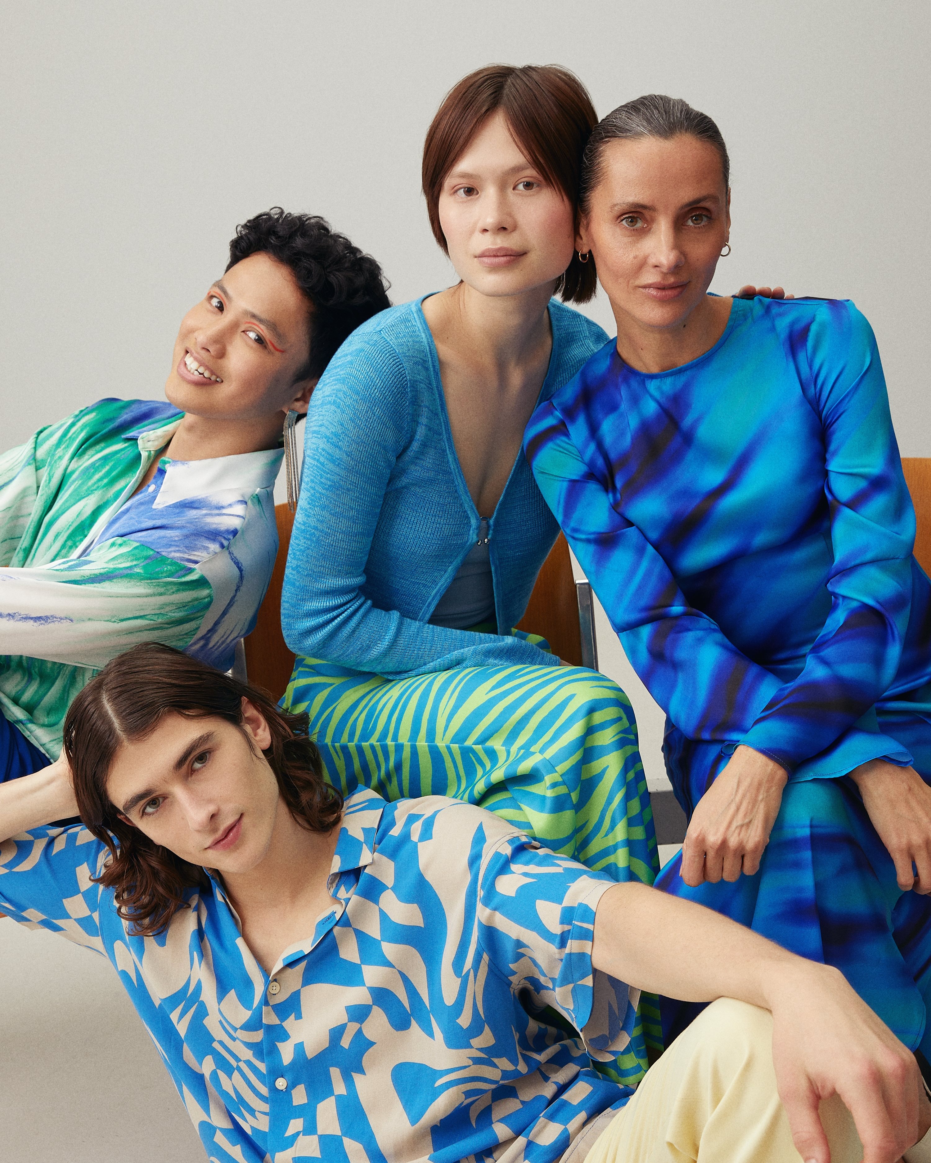 Four people posing closely together in a studio setting, wearing coordinated blue and green patterned outfits, conveying a modern, inclusive fashion aesthetic.