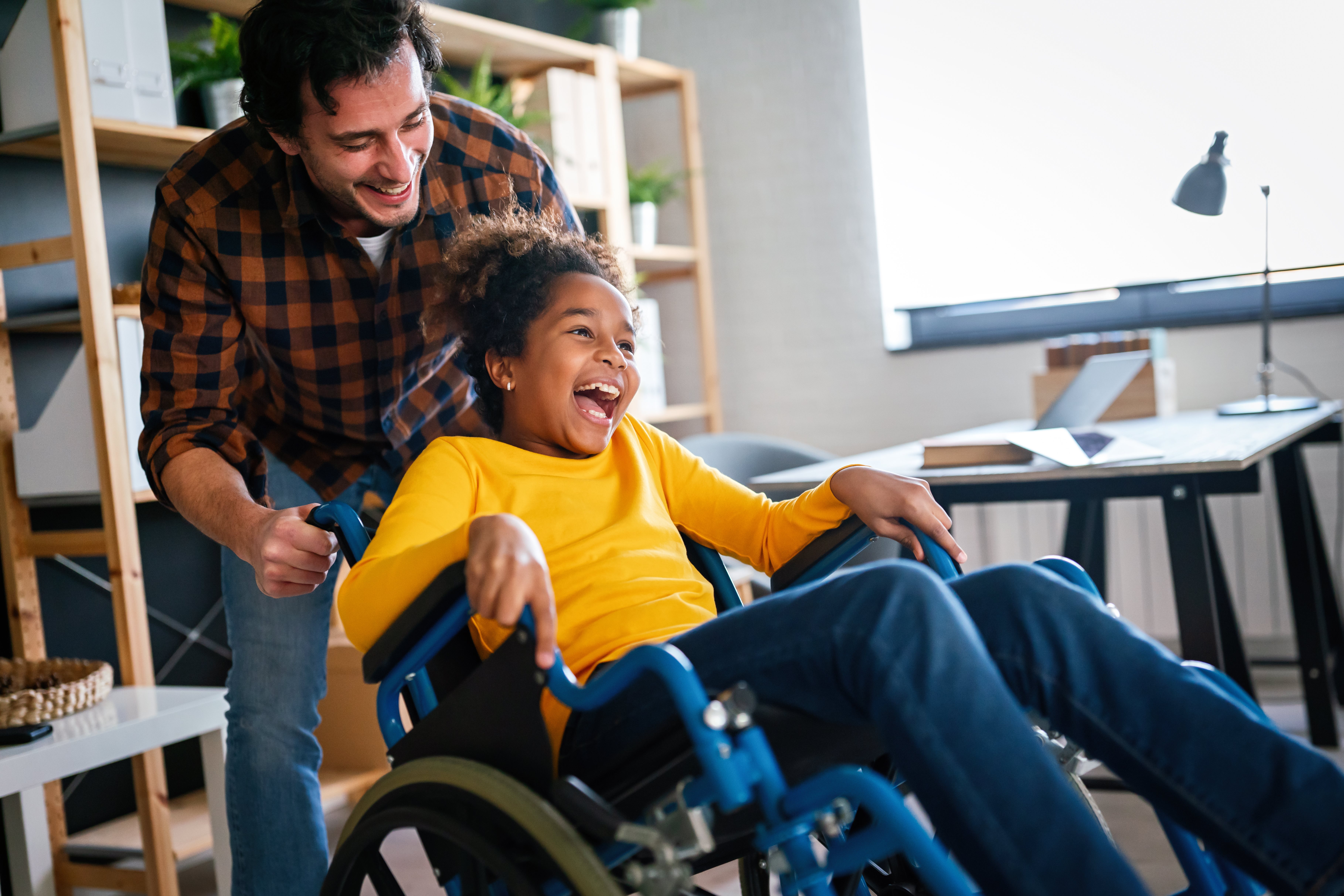 Adult pushing a smiling child in a wheelchair indoors, both laughing together in a bright home or classroom setting, conveying joy and inclusion.