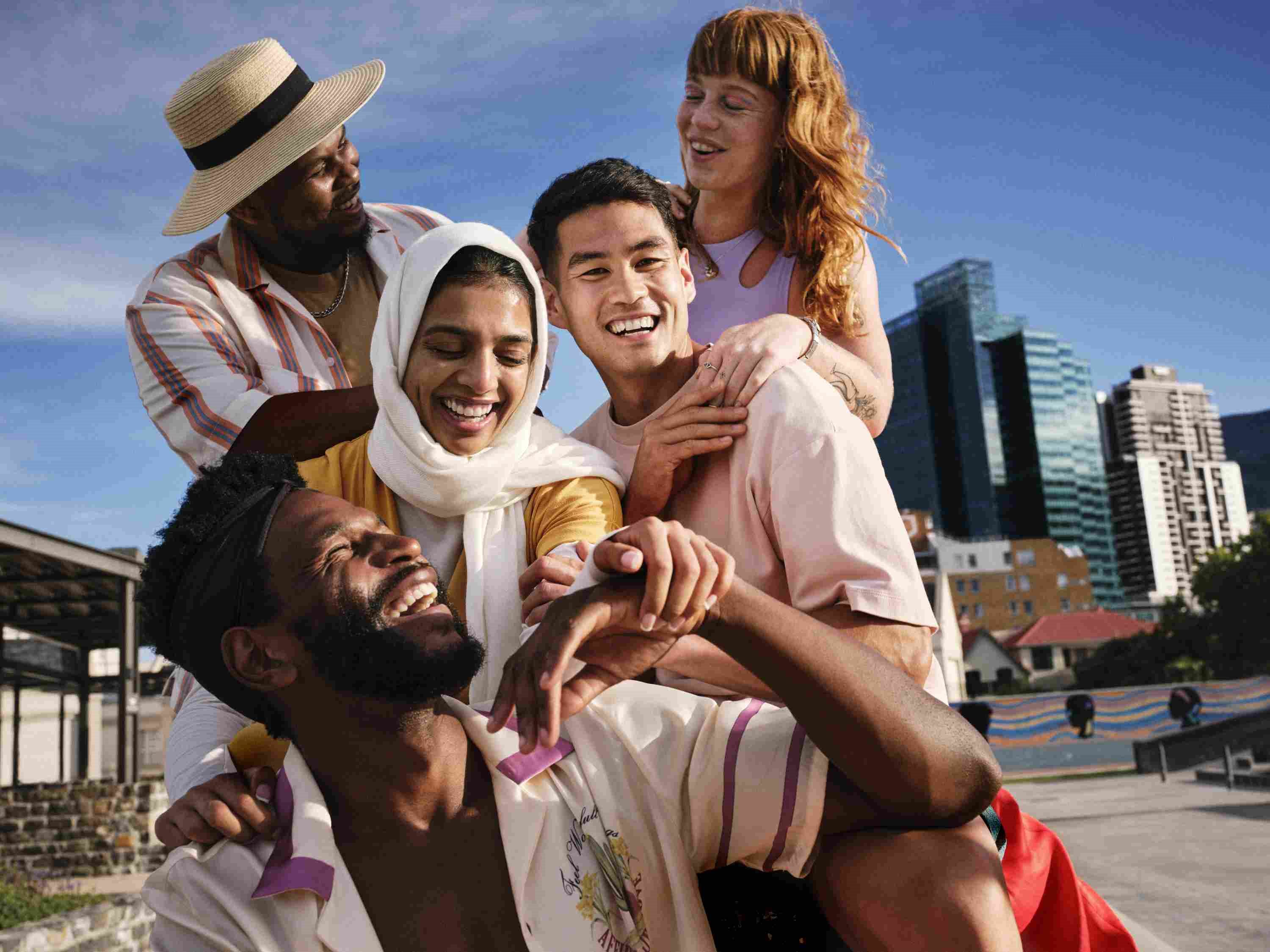 Group of five friends of diverse backgrounds laughing and posing together outdoors on a sunny day, showing close friendship and joy with a city setting in the background.