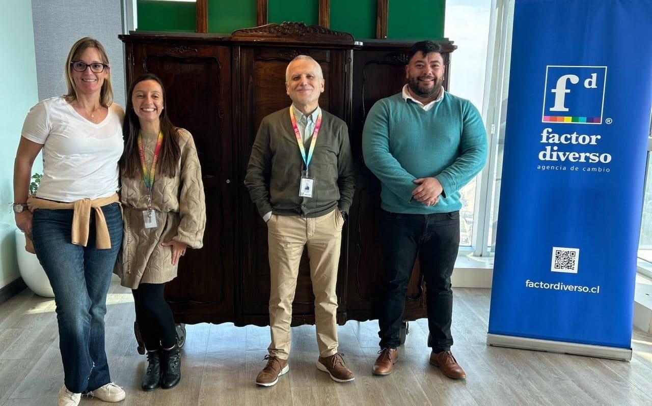 Four people standing indoors in front of a large wooden wardrobe, next to a blue banner with the logo and text “factor diverso” in Spanish. The setting is a modern office with large windows and hanging lamps.