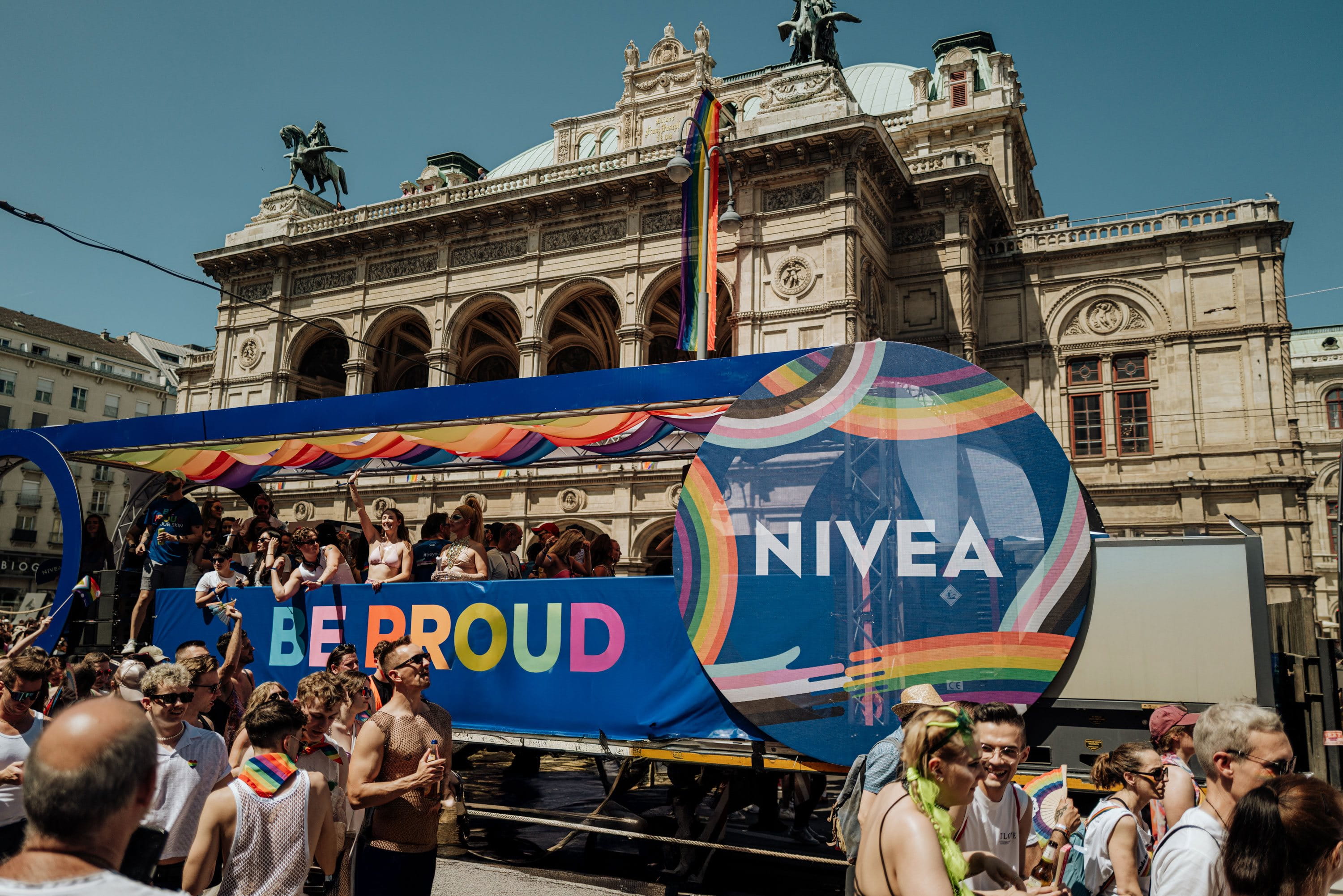 A colorful NIVEA-branded Pride parade truck with the slogan “BE PROUD” in rainbow colors, surrounded by people celebrating in front of a historic building with rainbow flags.