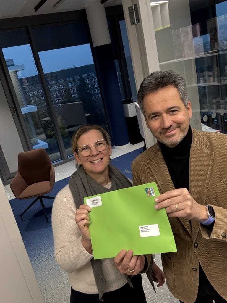 Two people standing indoors, smiling and holding up a bright green envelope together, with office windows and furniture in the background.