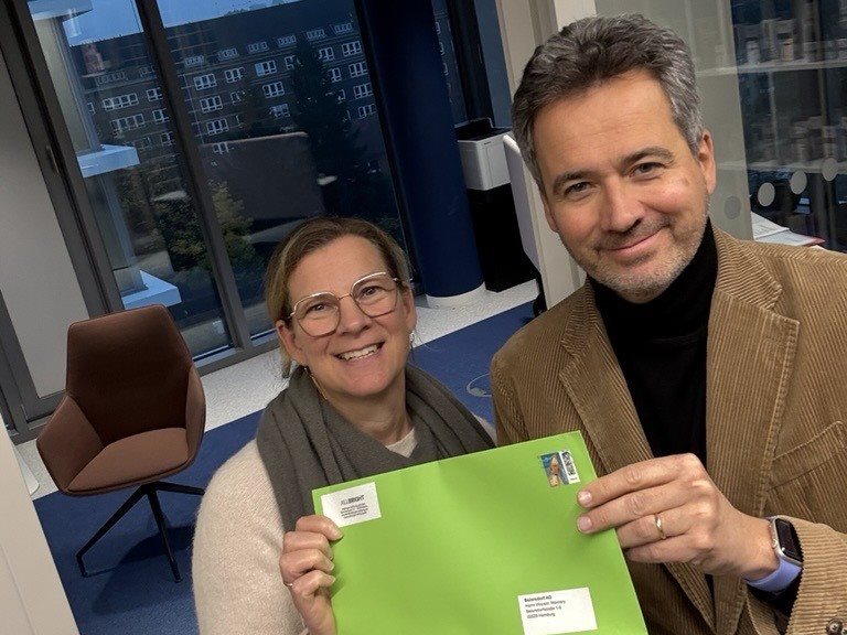 Two people standing indoors, smiling and holding up a bright green envelope together, with office windows and furniture in the background.