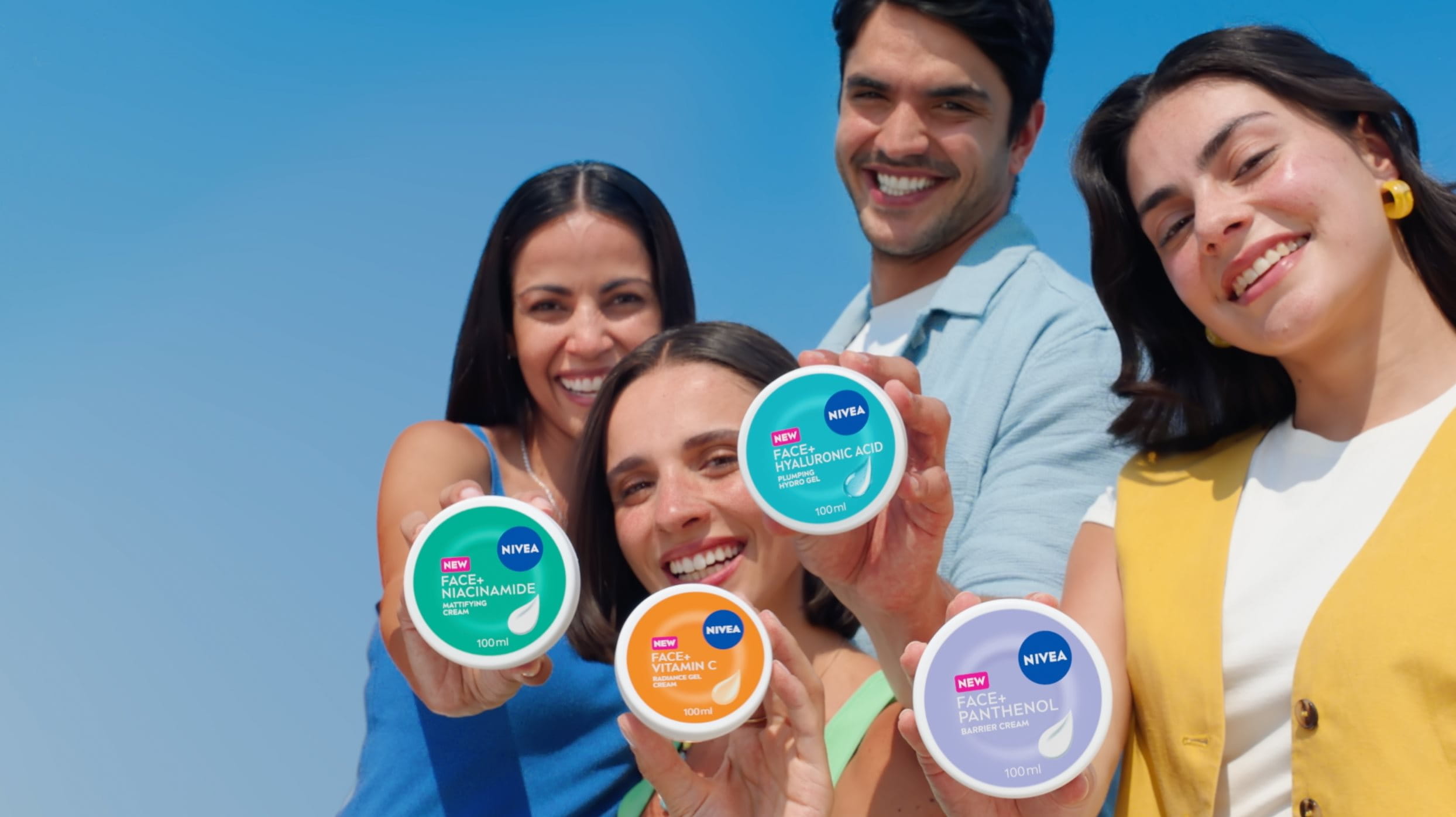 People smiling and showing NIVEA FACE+ cream jars in turquoise, orange, blue, and violet, each featuring the NIVEA FACE+ logo and a distinctive color-coded design.