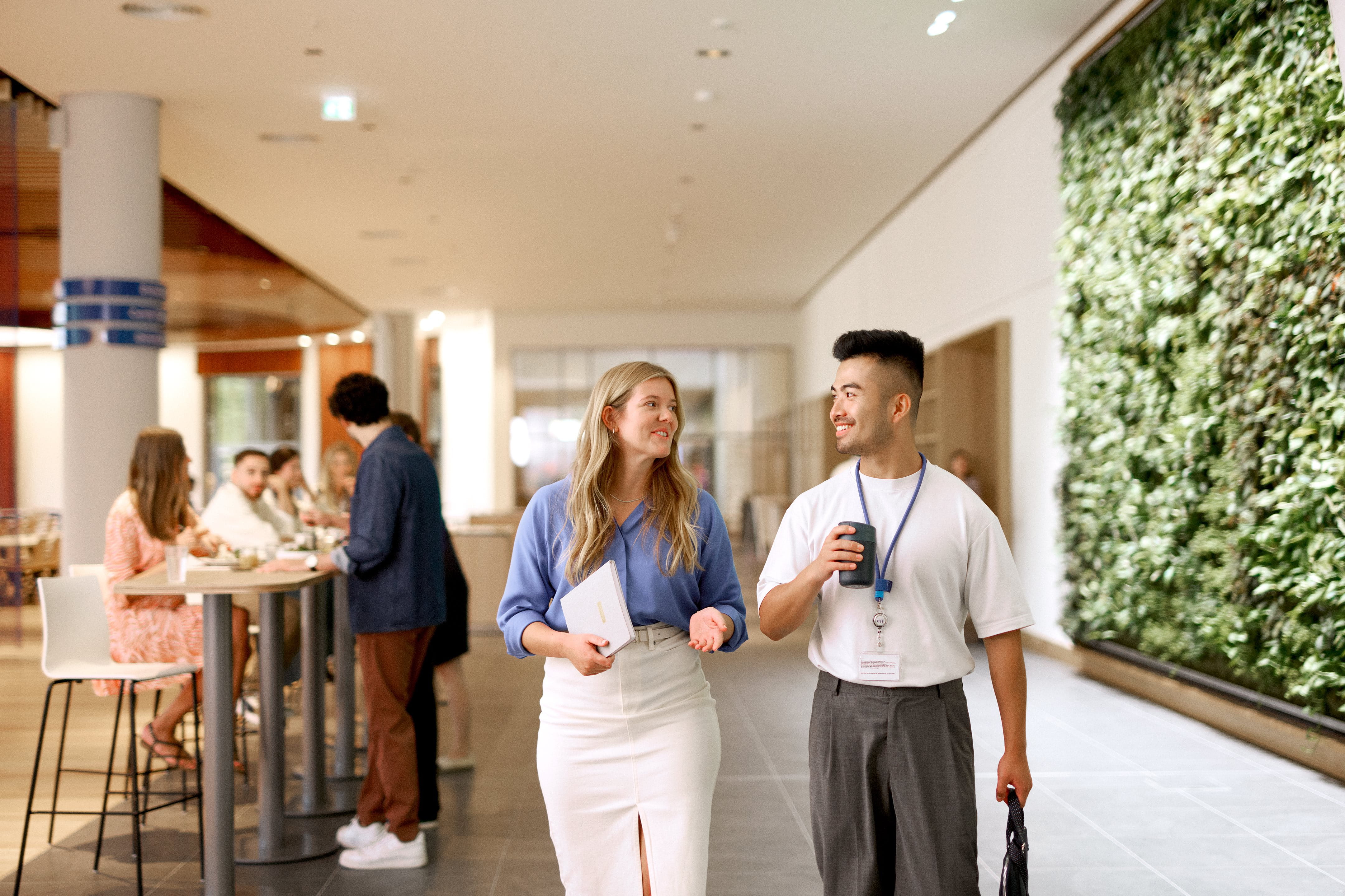 People standing and talking in a bright indoor hallway with plants on the wall, including two individuals in the foreground holding drinks and chatting while others converse in the background.