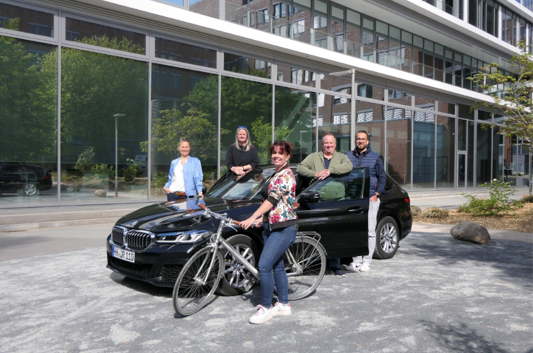 Group of people posing outdoors in front of a modern building, with one person standing beside a bicycle and others leaning against a black car.