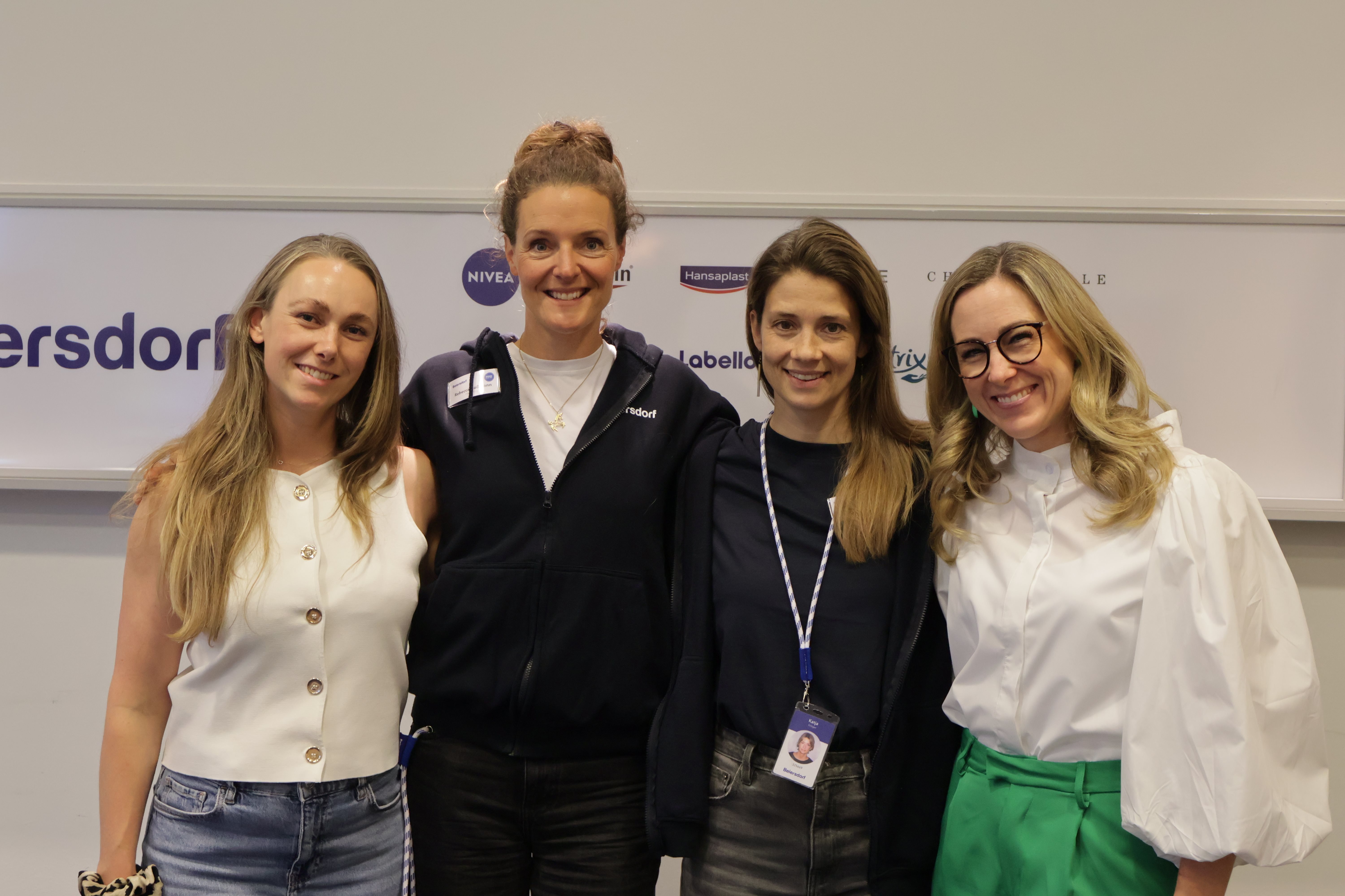 Four women standing side by side indoors at an event, smiling at the camera, with a visible sign in the background.
