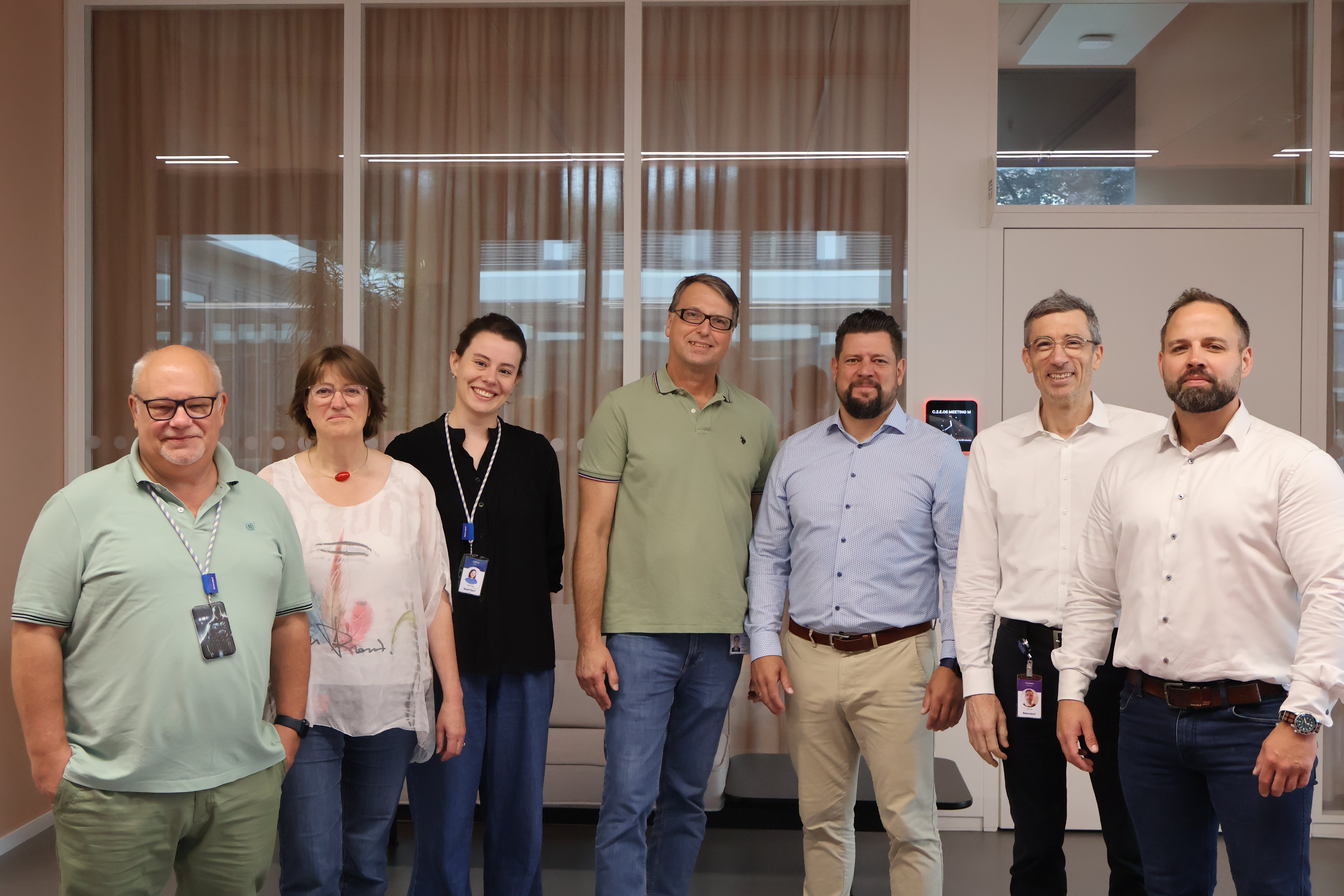 Group of colleagues standing indoors in an office setting, posing together and smiling, with glass walls in the background.