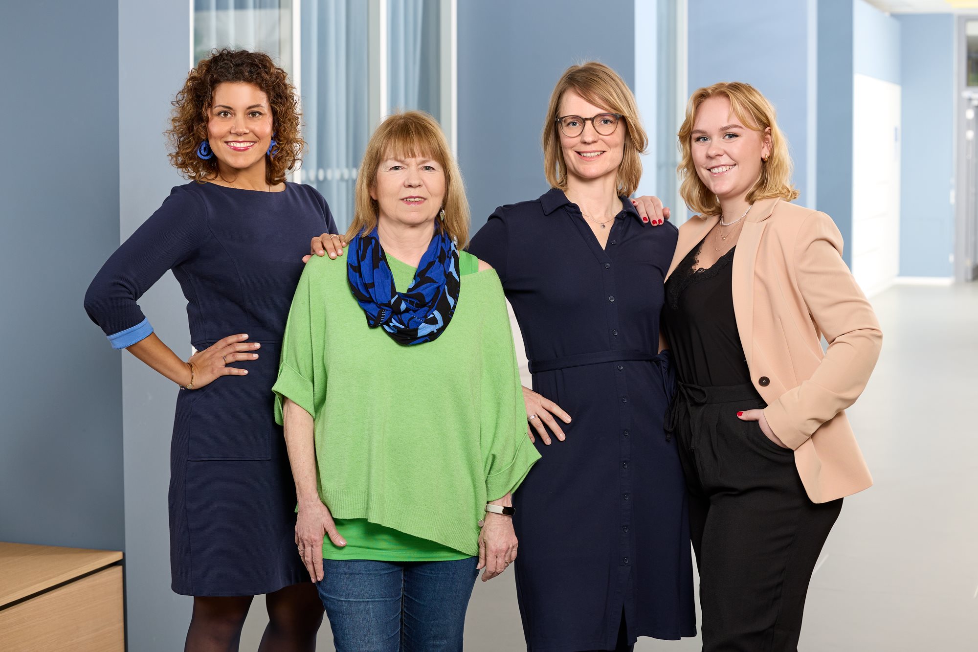 Four women standing together indoors, posing and smiling in front of tall windows, dressed in smart-casual and business attire.