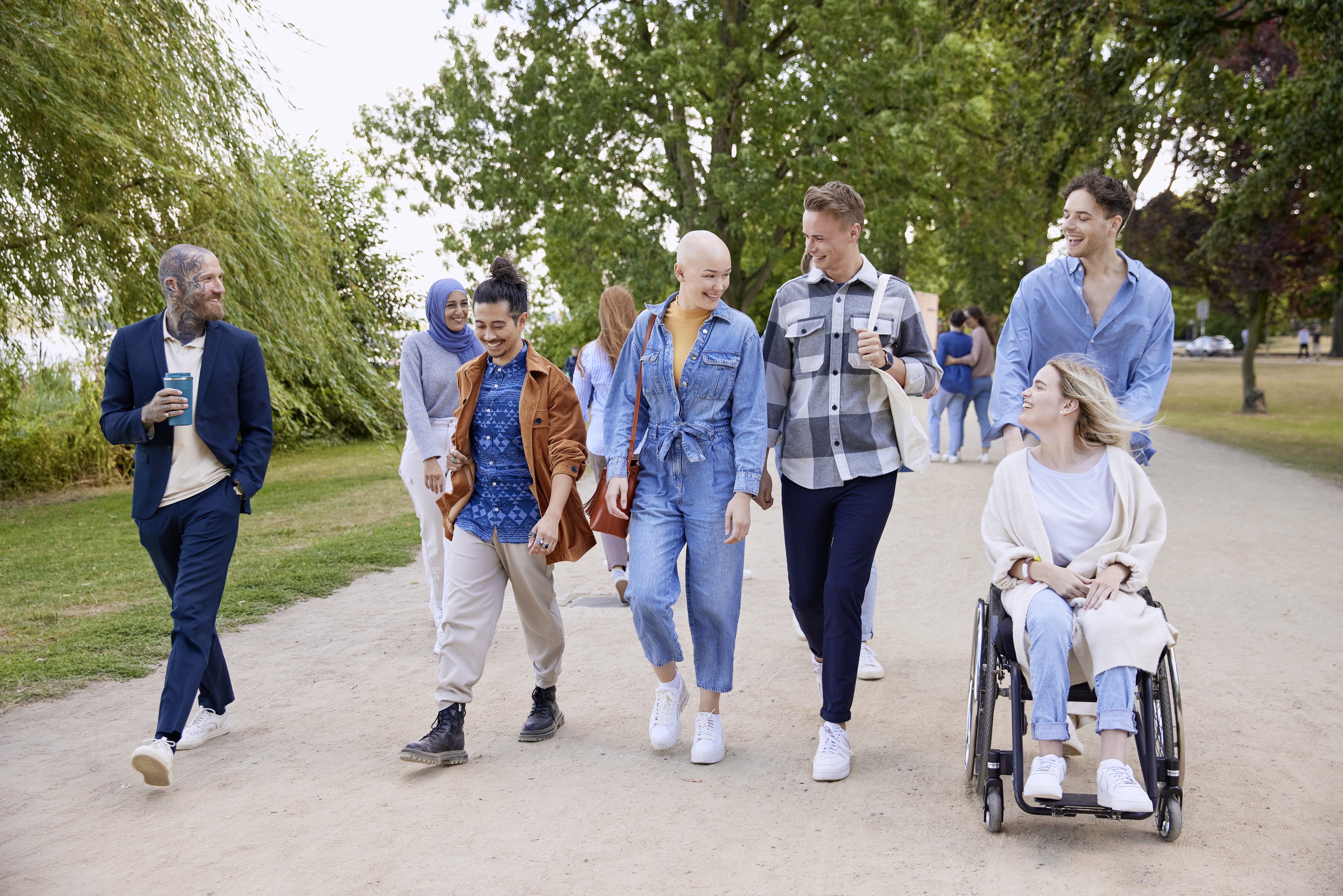 A group of people walking outdoors in a park-like setting, with a bald person in a blue denim outfit smiling while talking to others beside them.