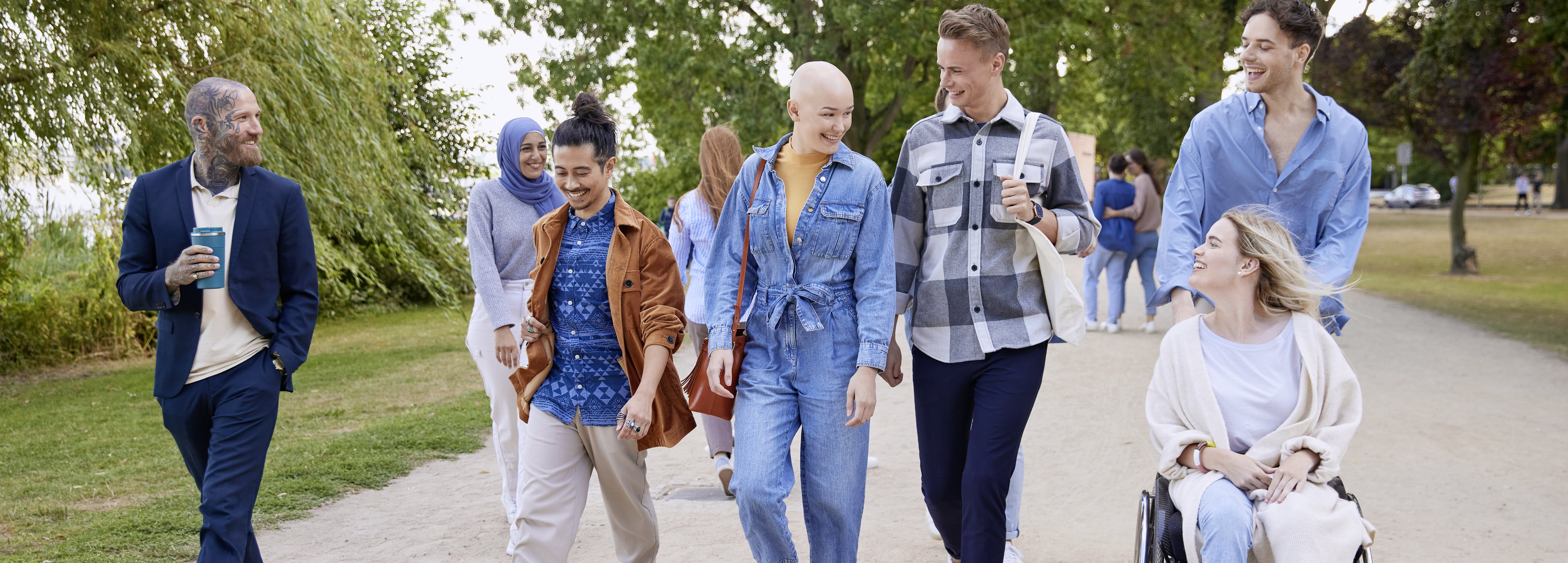 A group of people walking outdoors in a park-like setting, with a bald person in a blue denim outfit smiling while talking to others beside them.