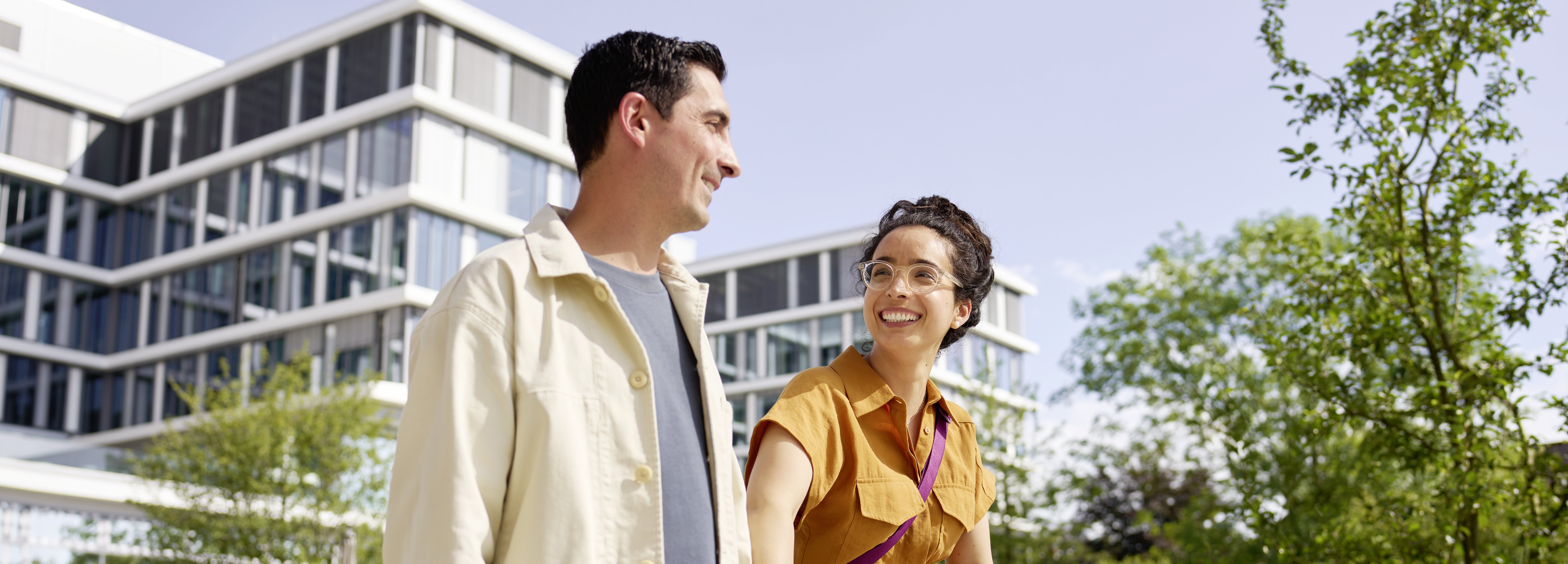 Two colleagues walking and talking outdoors on a modern office campus, with contemporary buildings and greenery in the background.