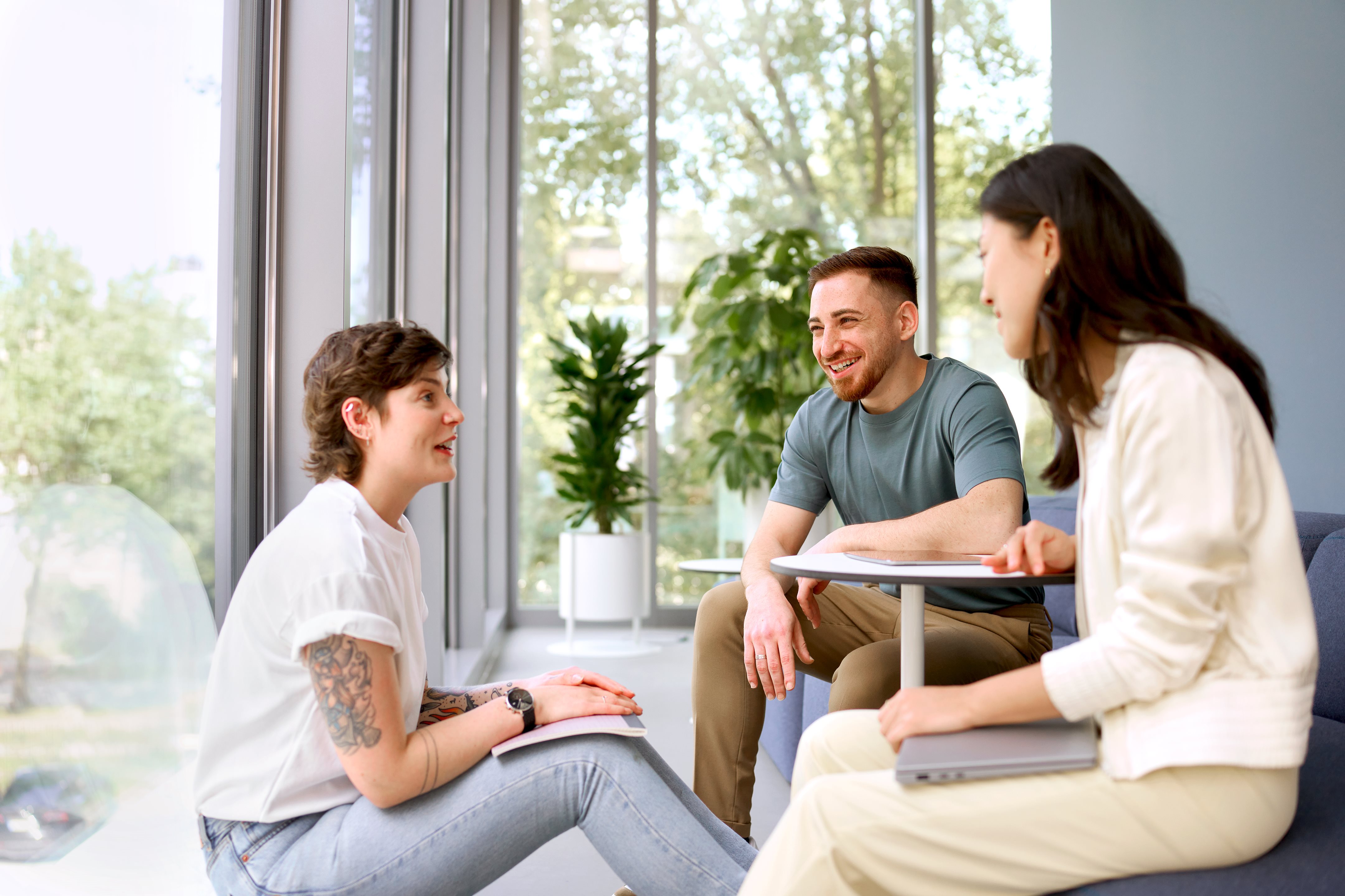 Three people seated in a bright room having a relaxed discussion, with two individuals listening while one person speaks during a small group meeting.