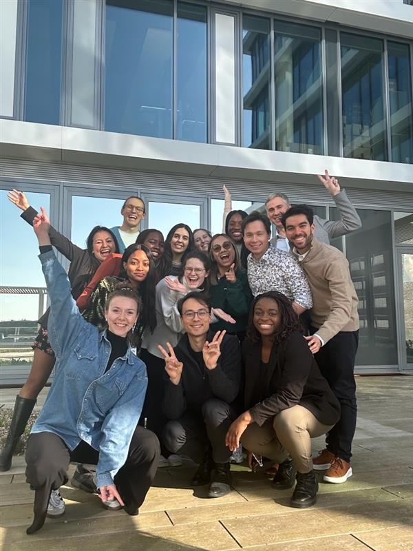 Group of colleagues posing together outdoors in front of a modern office building, smiling, cheering and making peace signs as a team photo.