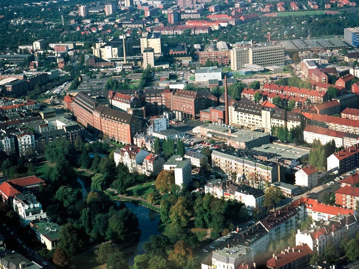 Aerial view of a historic urban campus with long red-brick buildings, modern extensions and tree-lined streets surrounded by city housing.