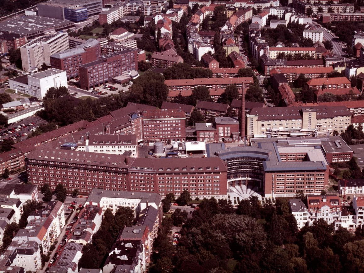 Aerial view of a large institutional complex with red-brick buildings, central courtyards and surrounding city streets, photographed from above.