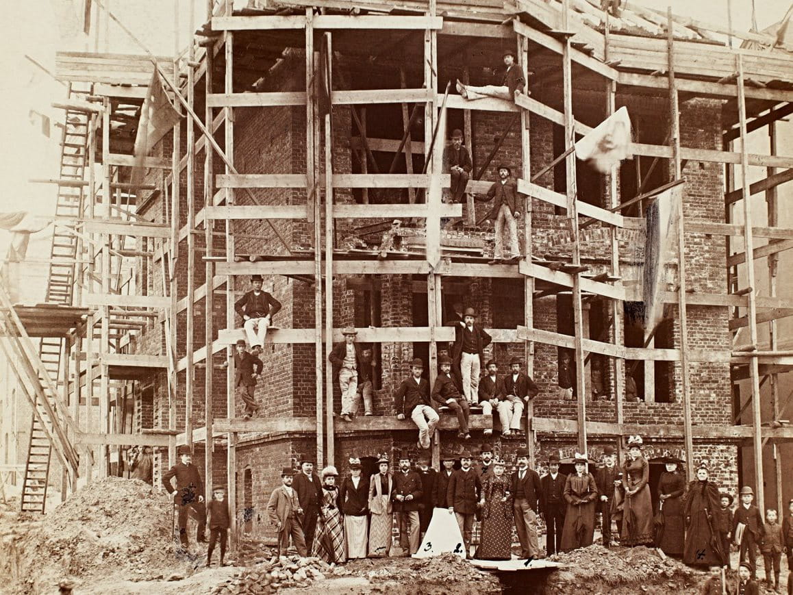 Historic black-and-white photograph showing a multi-storey building under construction, with scaffolding, exposed framework and groups of workers gathered at ground level.