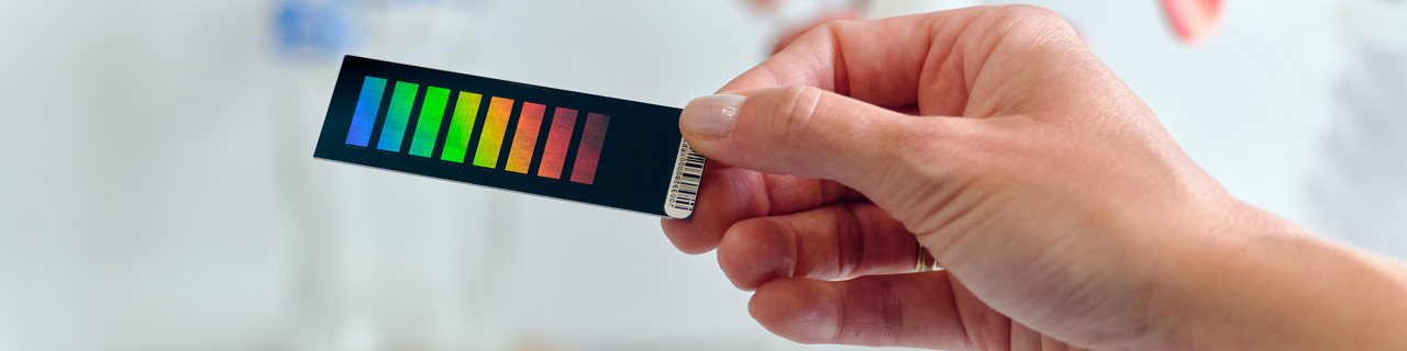 Hand holding a color test strip with multiple color indicators in front of laboratory equipment, suggesting a chemical or pH test in a lab setting.