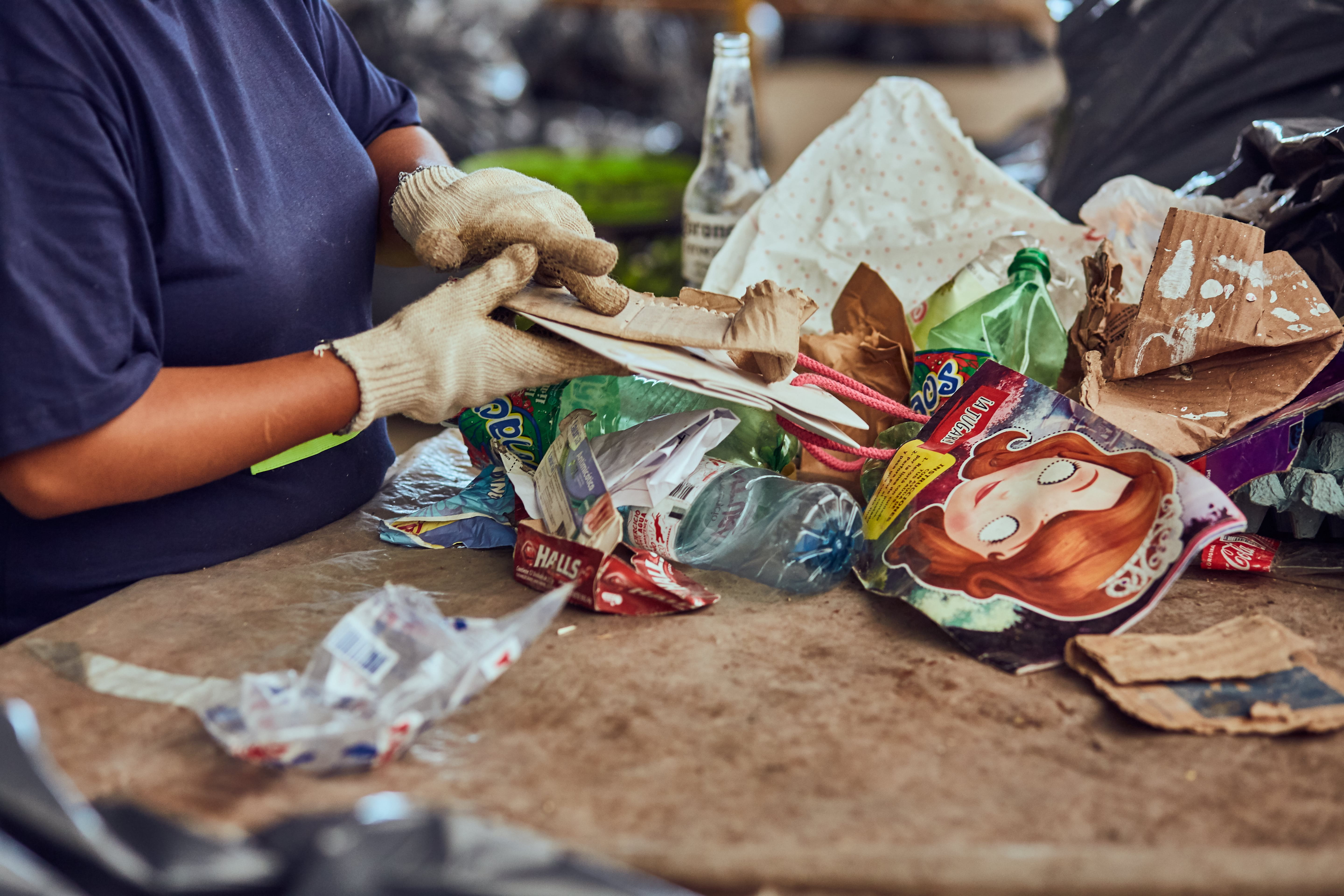 A person wearing gloves is sorting waste, including plastic bottles, cardboard, and packaging materials, on a table. The scene takes place indoors, where waste is being sorted for recycling or disposal.