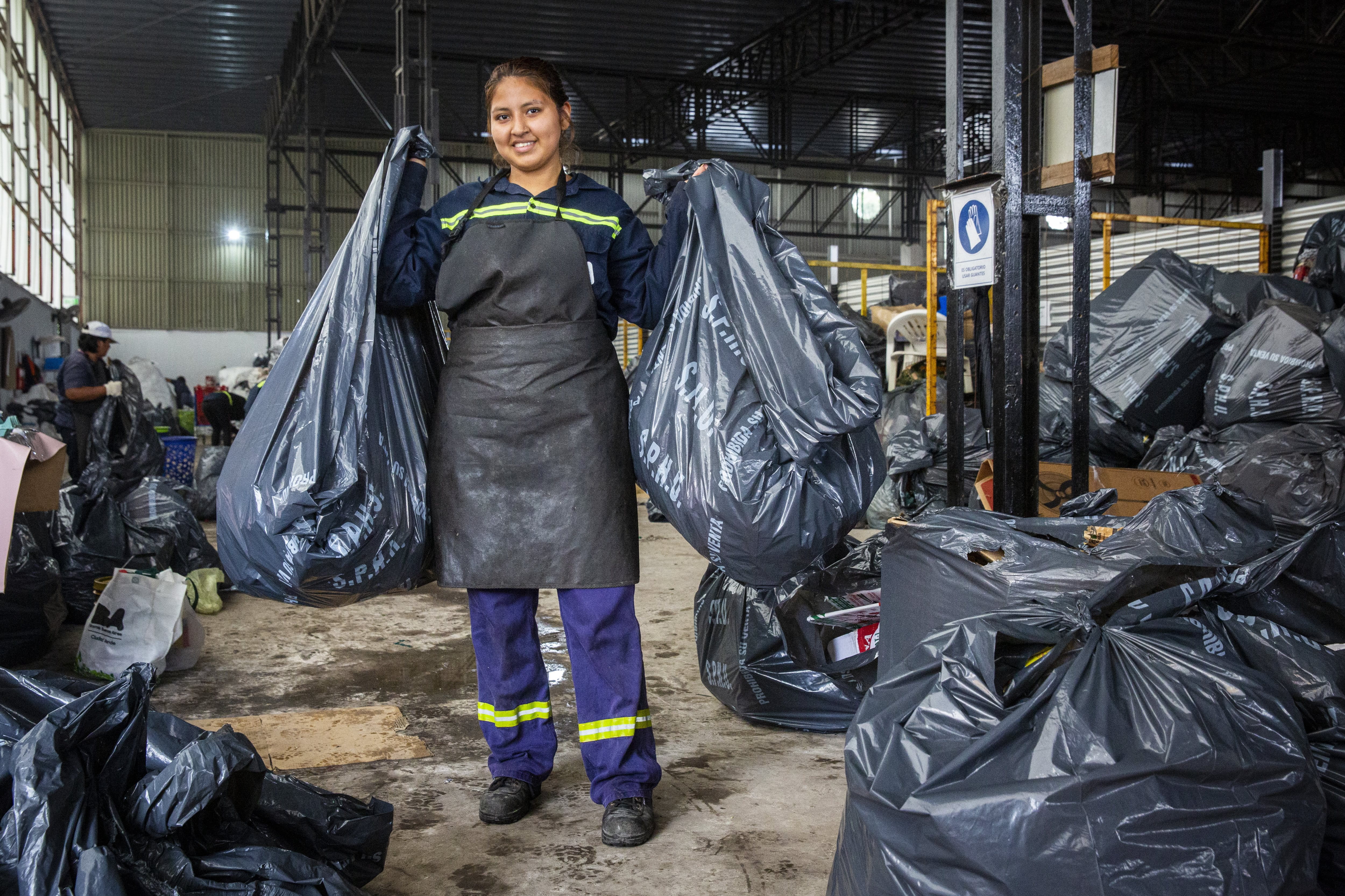 An Argentinian worker in protective clothing holds two large garbage bags in a warehouse in Argentina, surrounded by other collected waste. The scene takes place in an interior room where waste is sorted for recycling or disposal. 