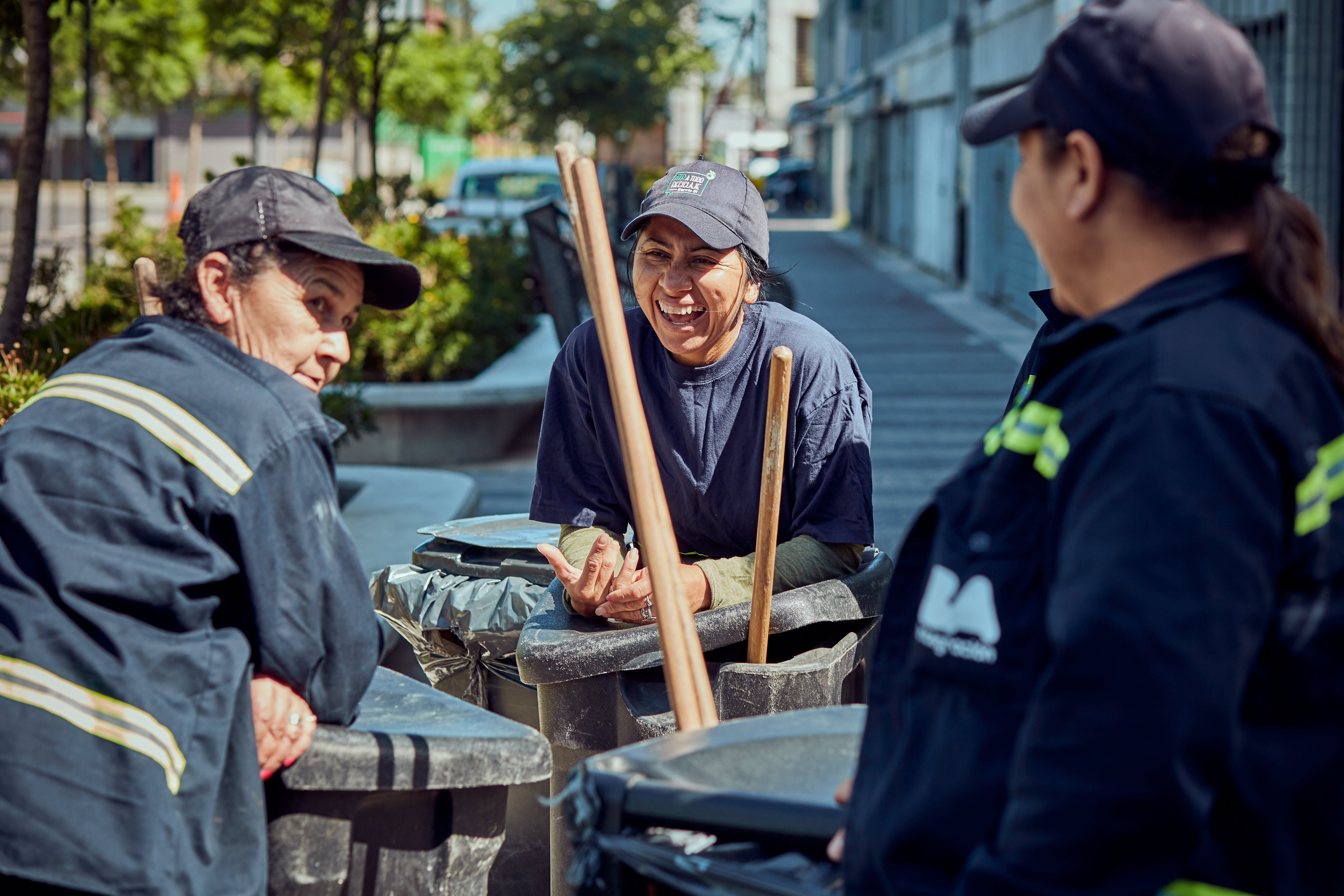 Three women in gray work clothes and hats stand together in a triangle on a sidewalk in front of a building. Each of the women is carrying a black, closed, waist-high garbage can, which has a kind of holder on the side with wooden handles sticking out of it. The picture suggests that the women are doing cleaning work on this street.
