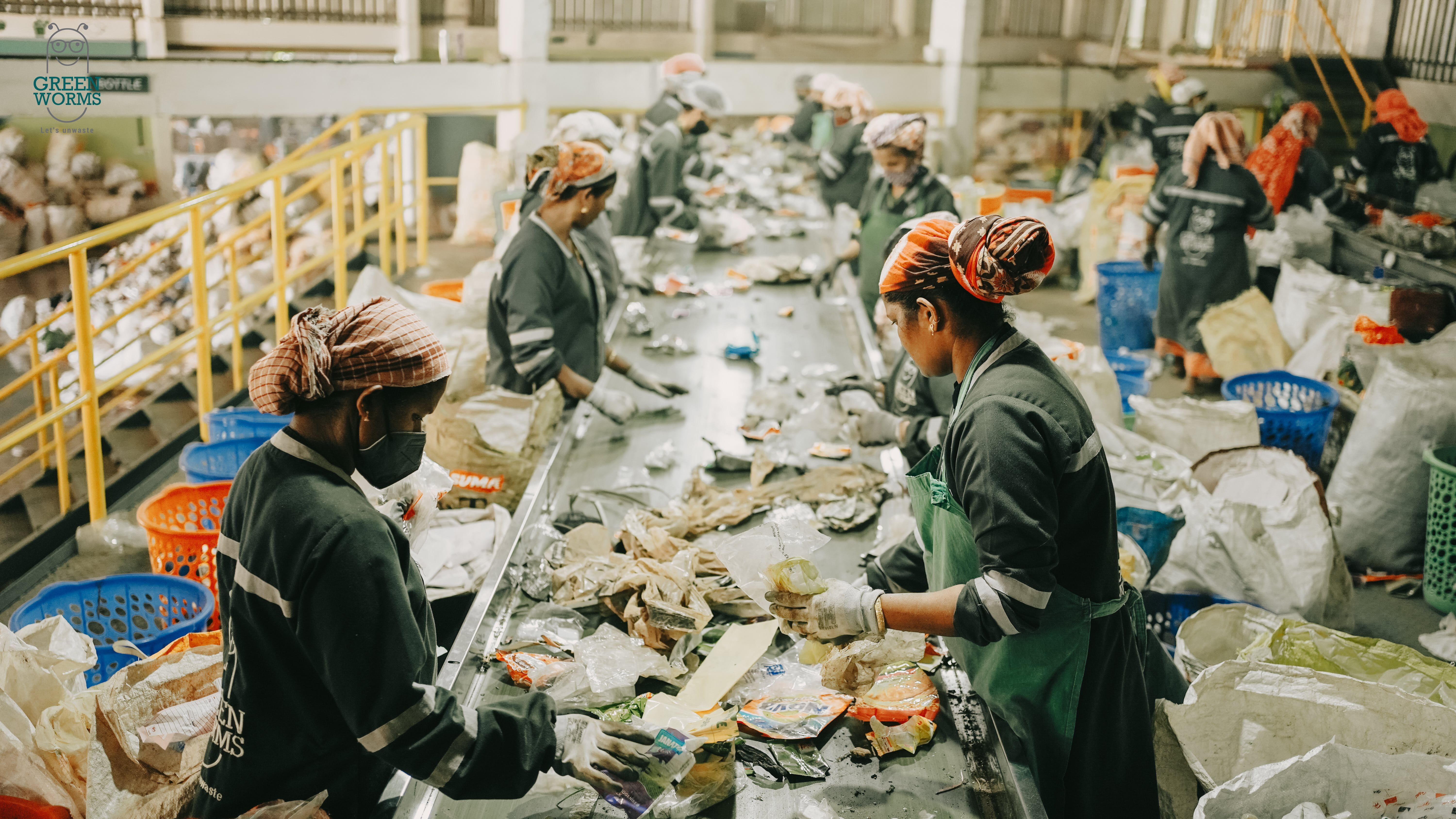 Workers sorting waste materials at a recycling plant in India. The workers are wearing protective clothing, including gloves and headgear. Various types of waste materials, including plastic and paper, can be seen on a long table. Next to the workers are baskets, presumably for sorting various recyclable materials. The recycling plant has a high ceiling with visible beams and lighting fixtures. A banner or sign with the text "GREEN WASTE WORKS" can be seen at the top left of the picture. 