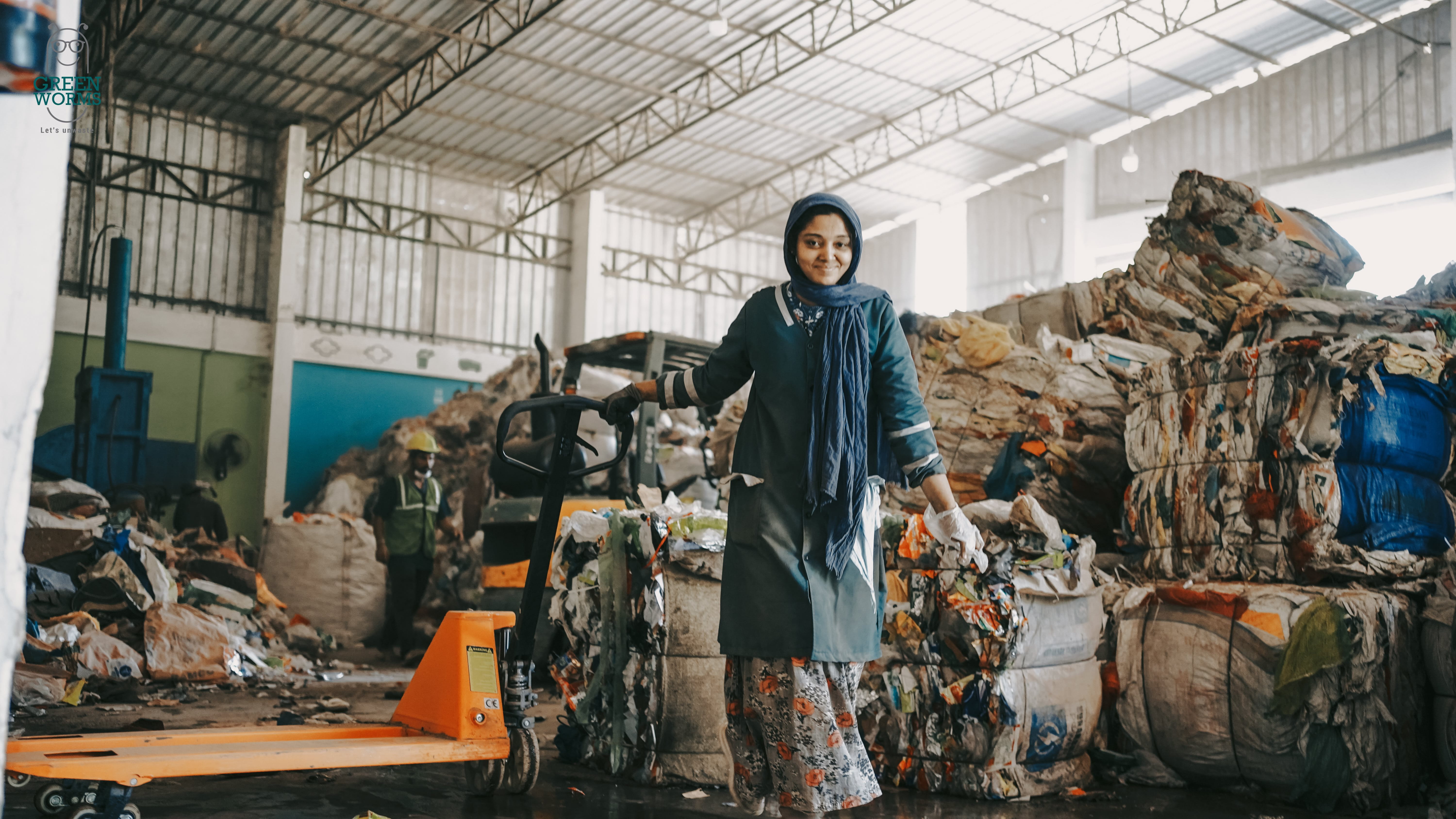 A Indian woman in a recycling facility in India stands next to an orange hand pallet truck. In the background, large bales of compressed recycled materials are visible. The scene takes place indoors, where waste is being sorted for recycling or disposal.