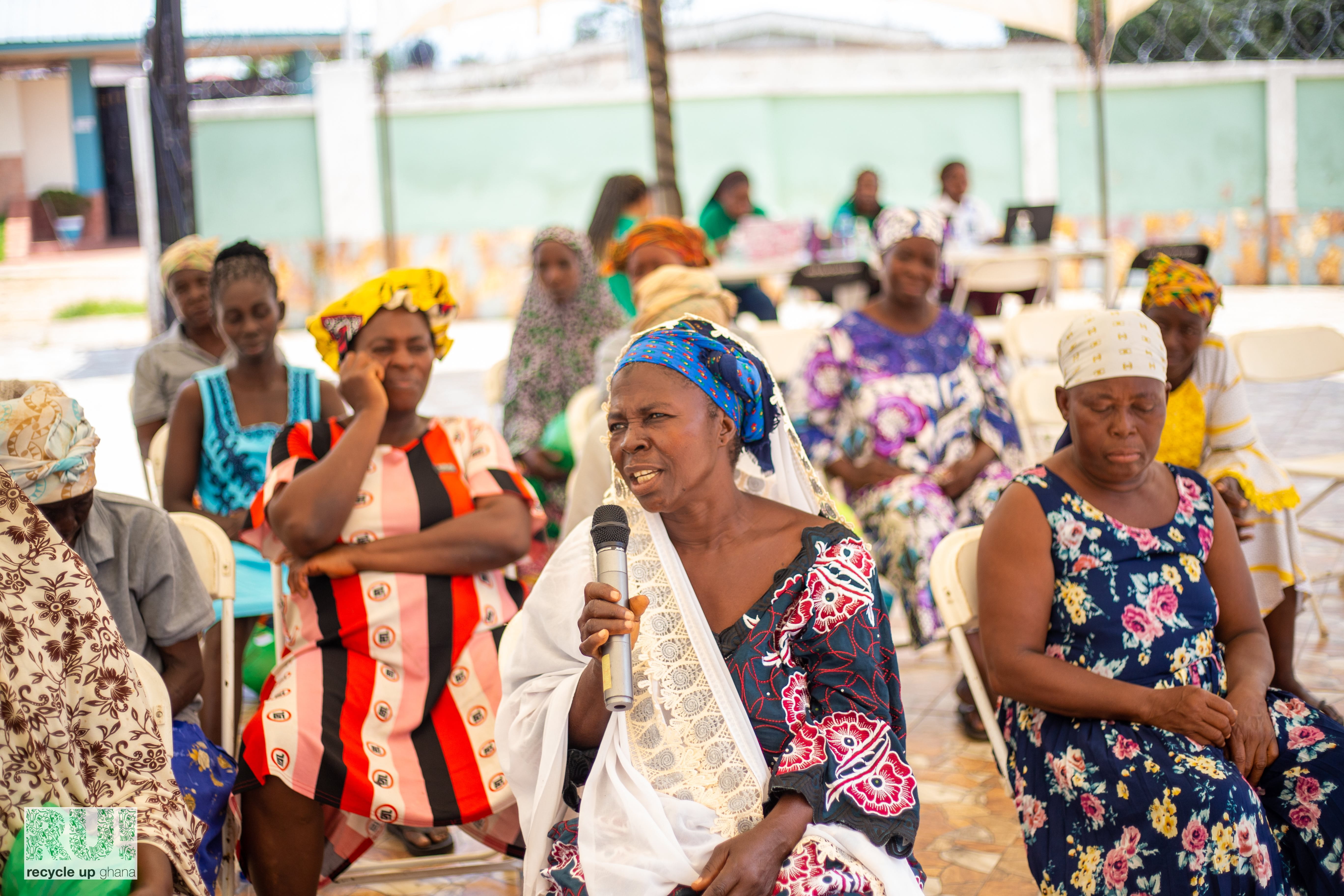 A group of dark-skinned women in colorful traditional dresses sitting on chairs outdoors. One person is holding a microphone, suggesting an event or meeting where people might be speaking or performing. 
