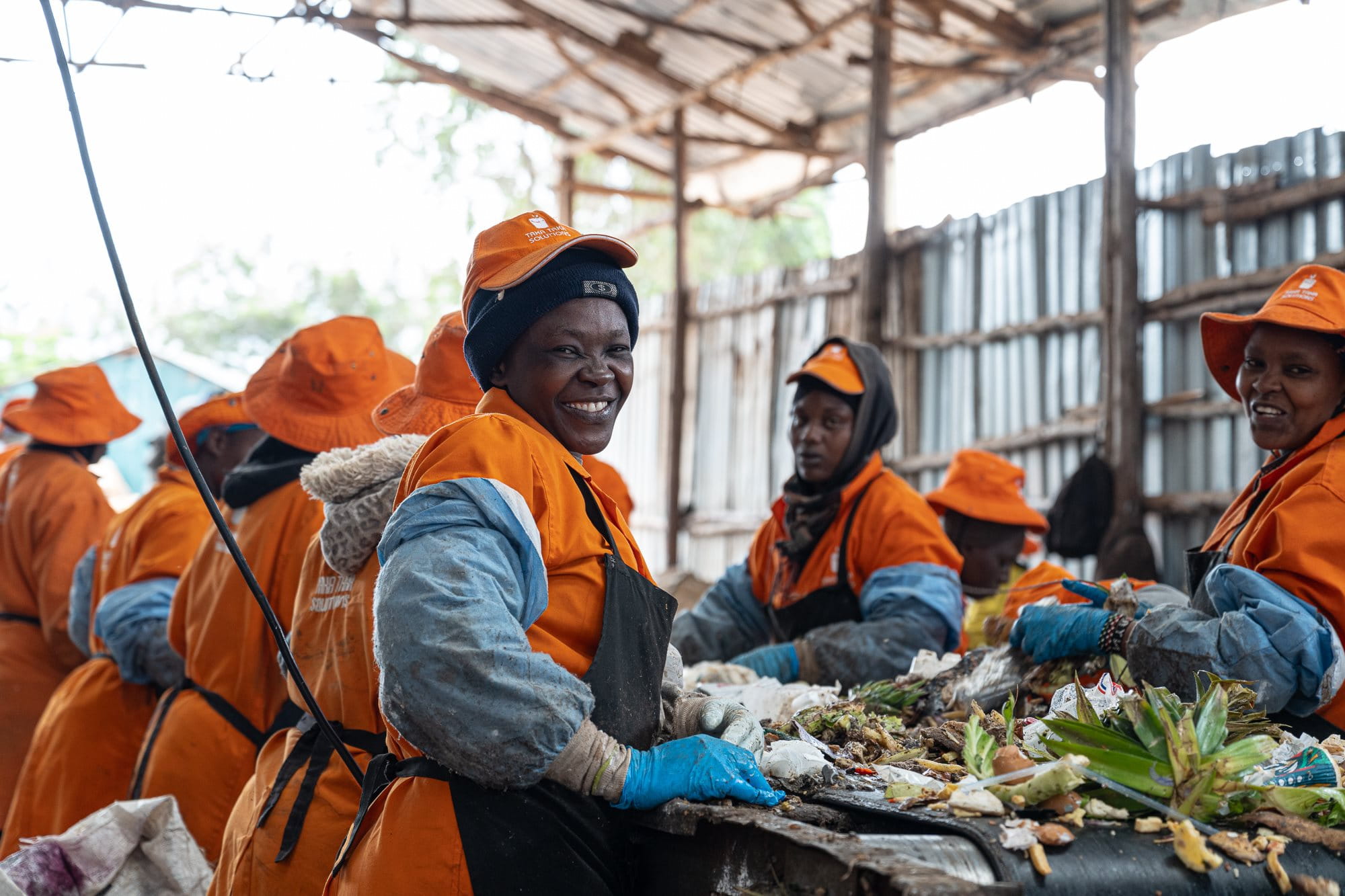 A group of workers in orange overalls and caps sorting waste at a recycling plant in Kenya. Under a corrugated iron roof supported by wooden beams, they are busy separating different types of waste materials on a long table. Plastic and organic materials are visible on the table. 