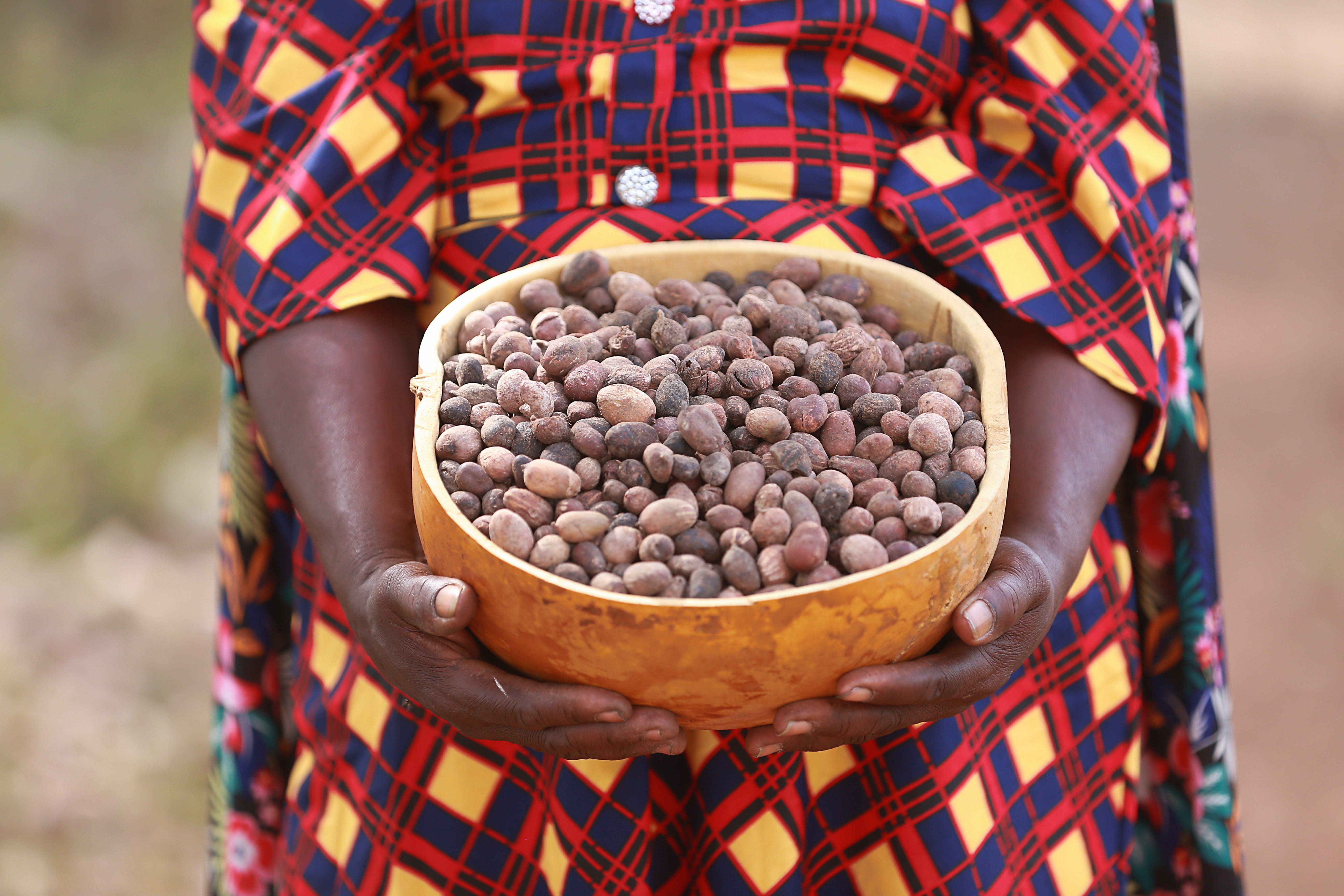 A medium shot of a smiling woman wearing a vibrant green and pink patterned dress and a white headscarf, sitting outdoors and waving at the camera. In the foreground is a large, rounded traditional clay stove, and in the background, a village scene with a thatched-roof hut is visible under a bright sky.