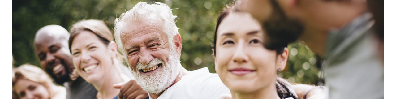 A close-up, outdoor portrait of a smiling elderly man with white hair and a white beard. He is wearing a white shirt and is flanked by the partial faces of two younger women, with a soft, out-of-focus green background.