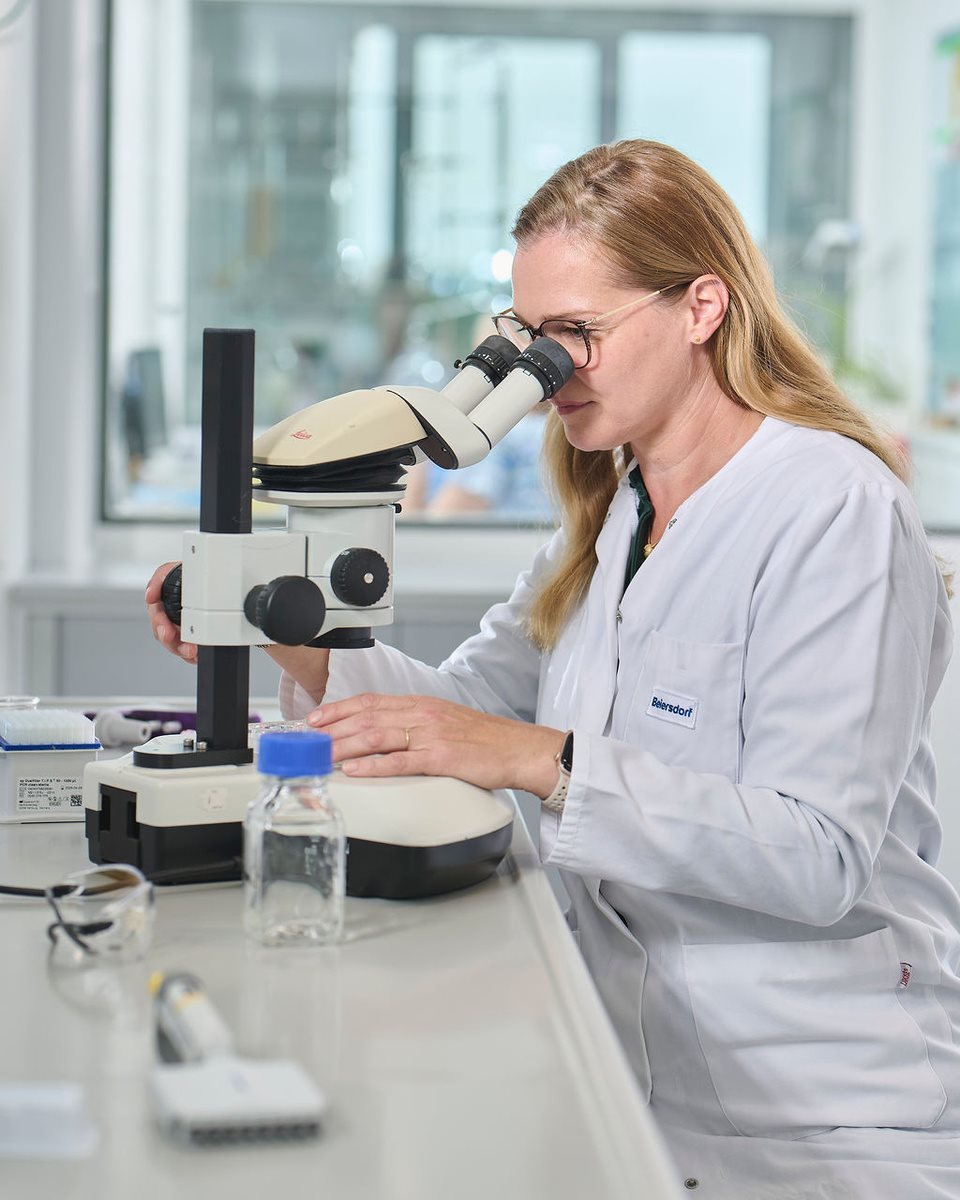 Female scientist wearing safety glasses using a microscope in a laboratory, with lab equipment and samples arranged on the workbench.