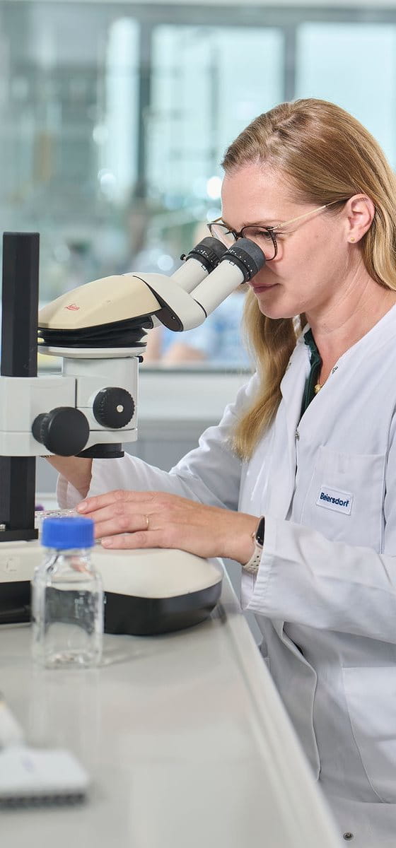 Female scientist wearing safety glasses using a microscope in a laboratory, with lab equipment and samples arranged on the workbench.