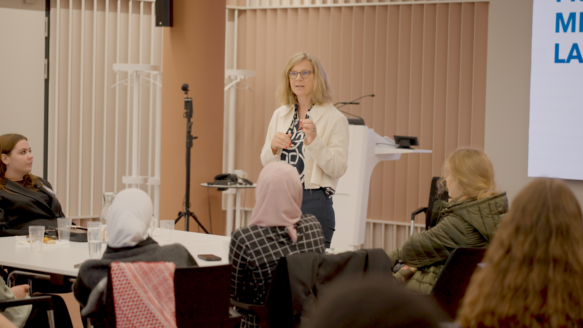 A woman speaking to a seated audience during a workshop or presentation in a modern indoor space, with attendees listening attentively.