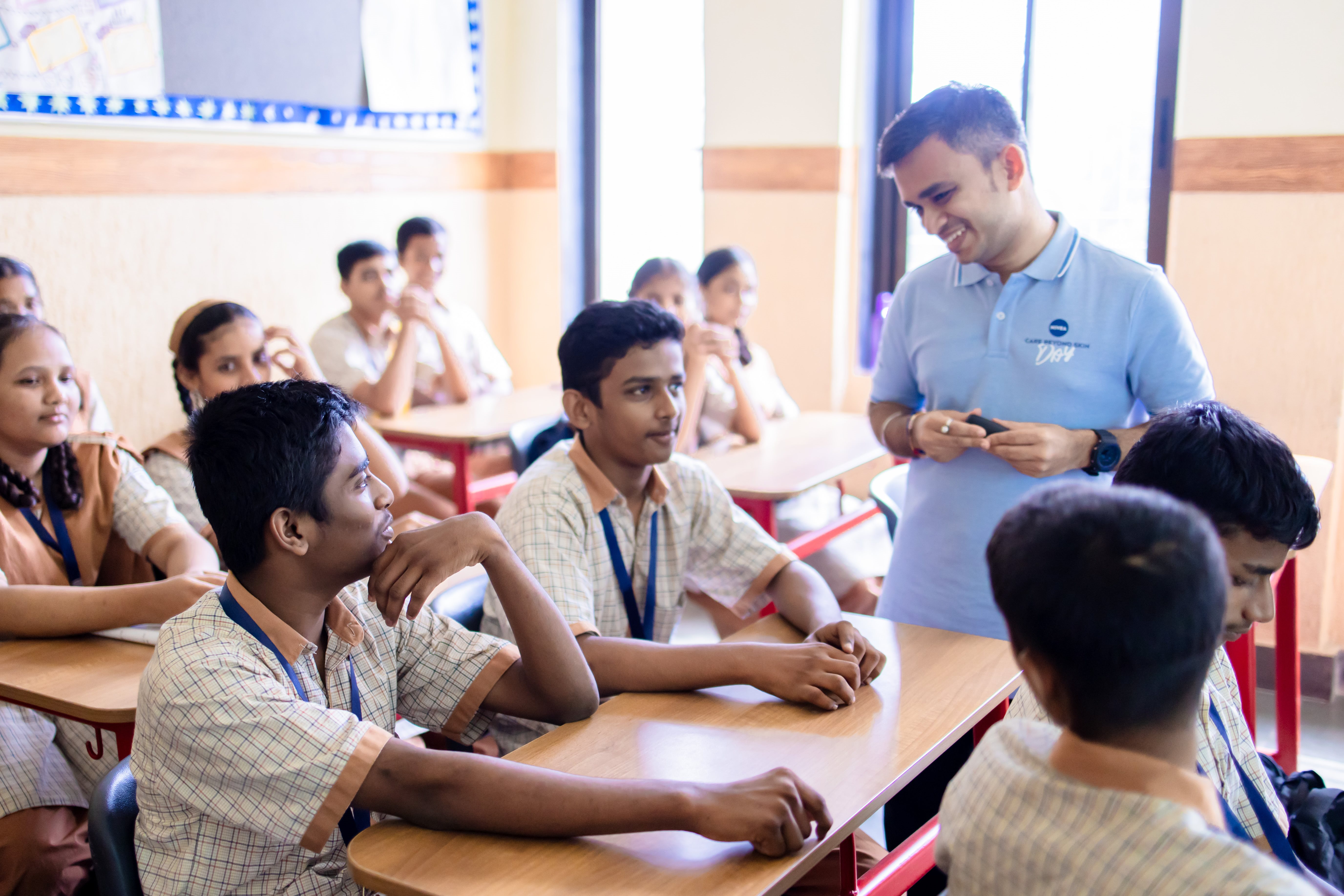 A person in a blue NIVEA shirt standing next to a group of people in classroom smiling and talking to them during CARE BEYOND SKIN Day.
