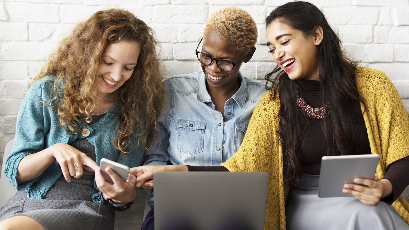 Three people sitting closely together indoors, smiling and laughing while looking down at something shared, conveying friendship and connection.
