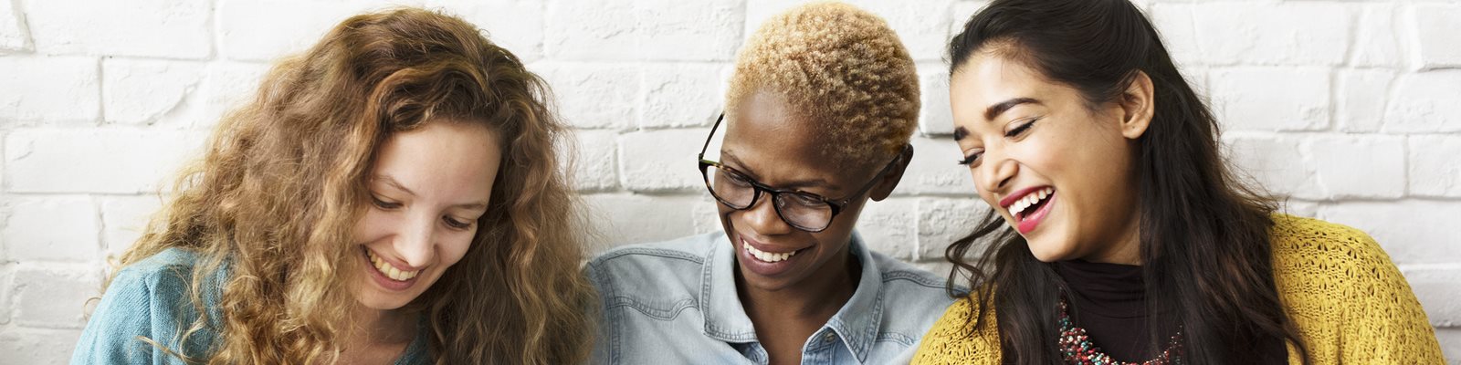 Three people sitting closely together indoors, smiling and laughing while looking down at something shared, conveying friendship and connection.