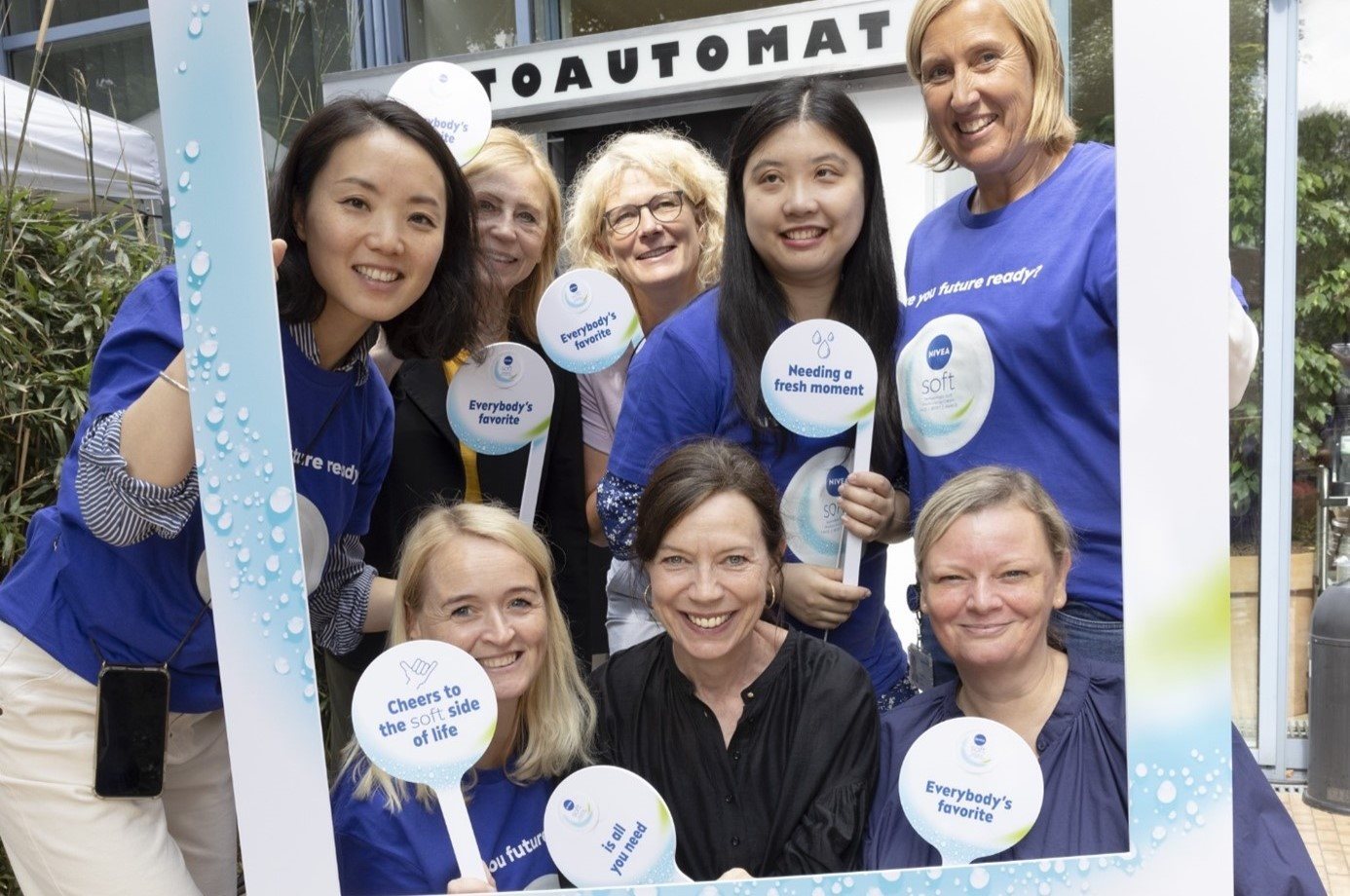 Group of smiling colleagues posing together at an outdoor event, holding branded signs and standing behind a decorative photo frame.