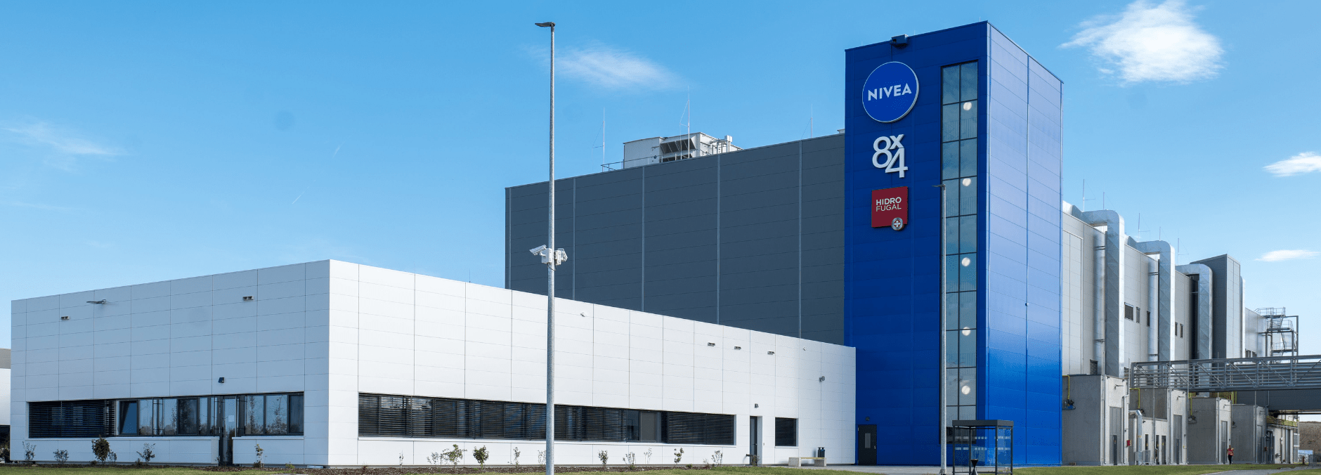 Exterior view of a modern industrial building with a grey and white facade, rooftop equipment, and tall flagpoles in the foreground under a clear blue sky.