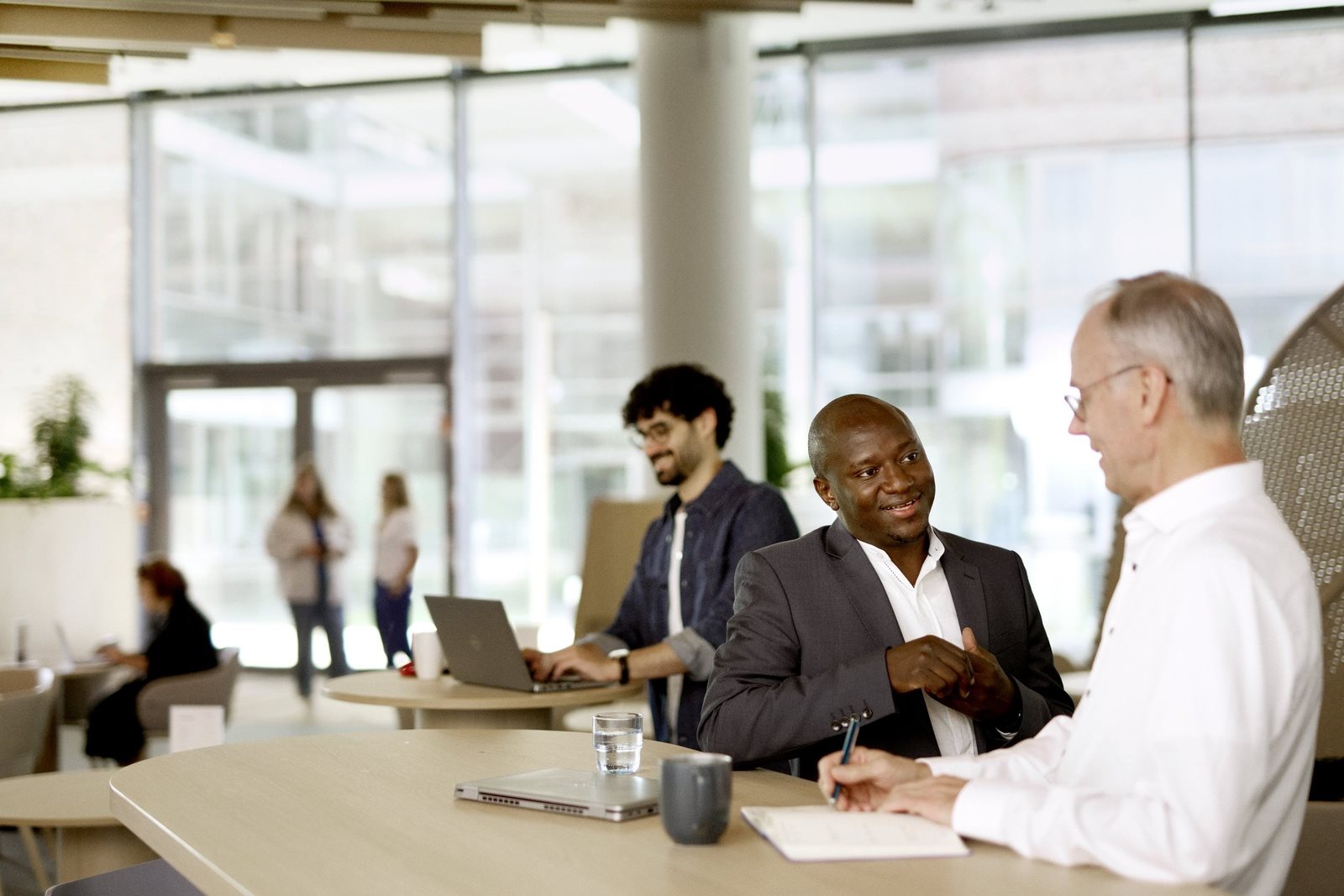 Three people sitting at a long table in a modern office or café space, talking and working, with laptops, notebooks, and drinks on the table.