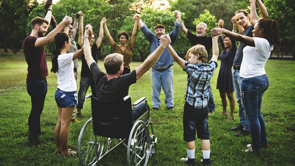 A diverse group of people standing in a circle on a grassy field, holding hands; one person using a wheelchair is included in the circle.
