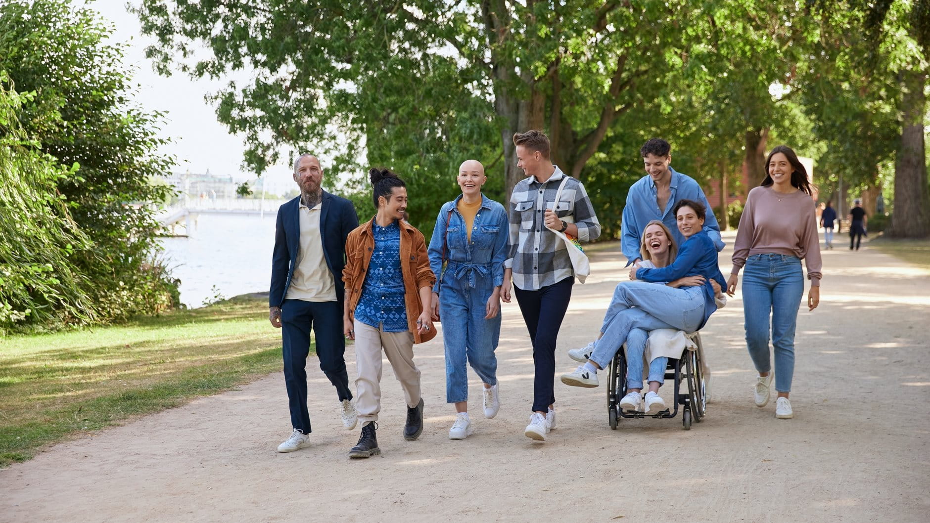 Group of people walking together outdoors along a tree-lined path, smiling and talking, with one person using a wheelchair.