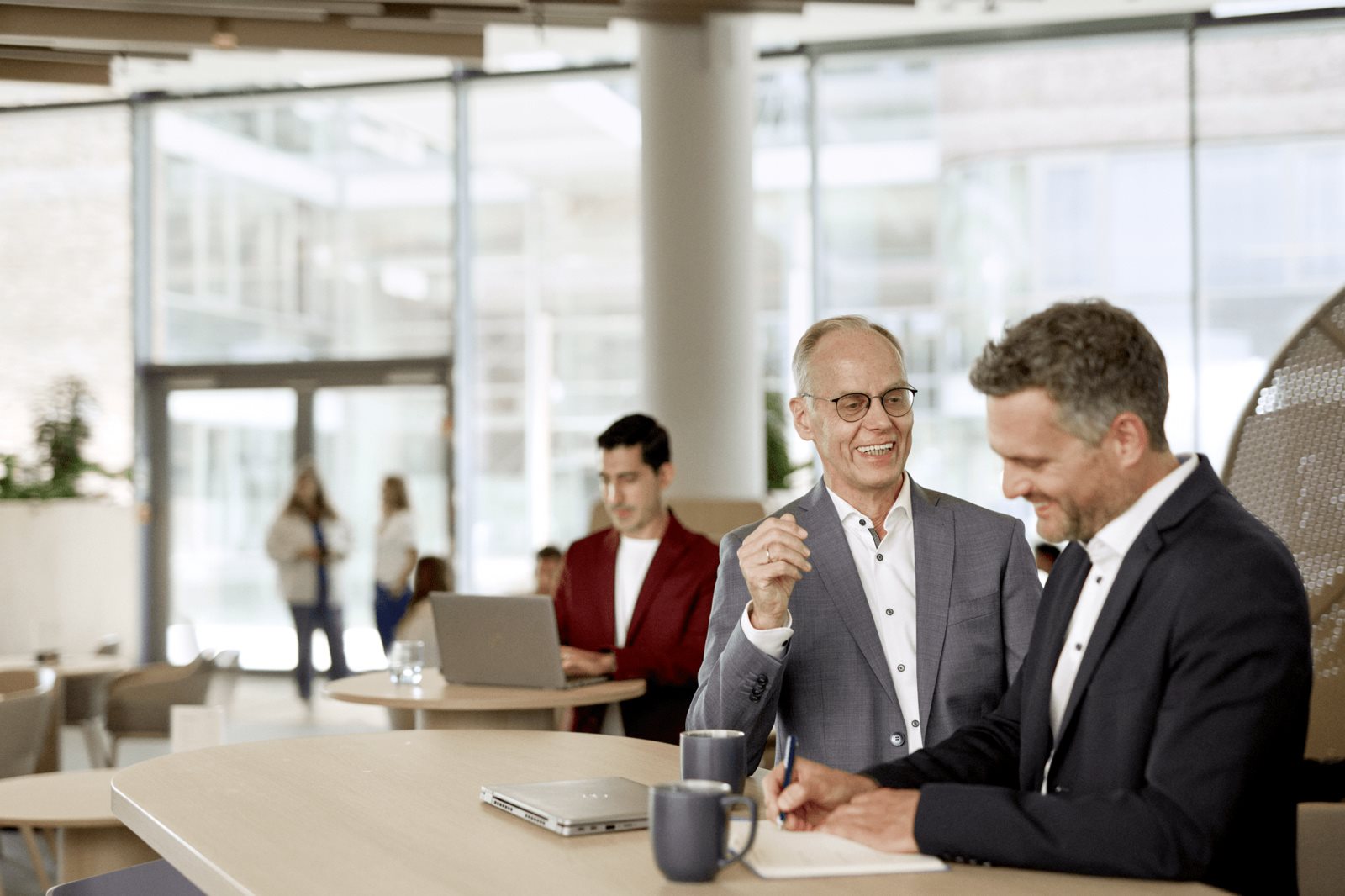 Three business professionals in suits talking together in a modern office lobby, with one person gesturing while the others listen.