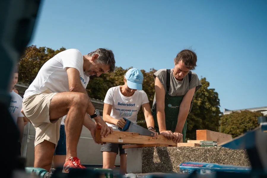 “Three people working together outdoors on a woodworking task, measuring and securing wooden planks on a workbench under a clear blue sky.