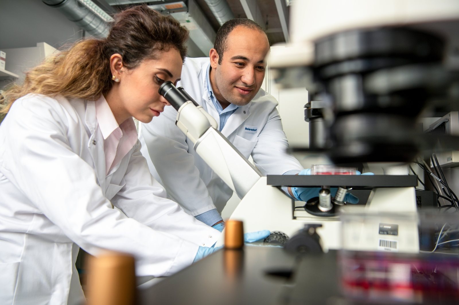 Two scientists in lab coats working together in a laboratory, with one looking through a microscope while the other observes and discusses the results.