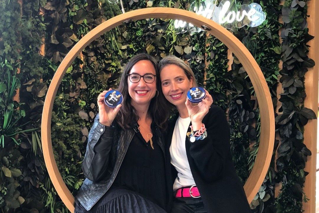 Two women smiling and holding small cosmetic jars while posing together in front of a decorative circular frame with greenery.